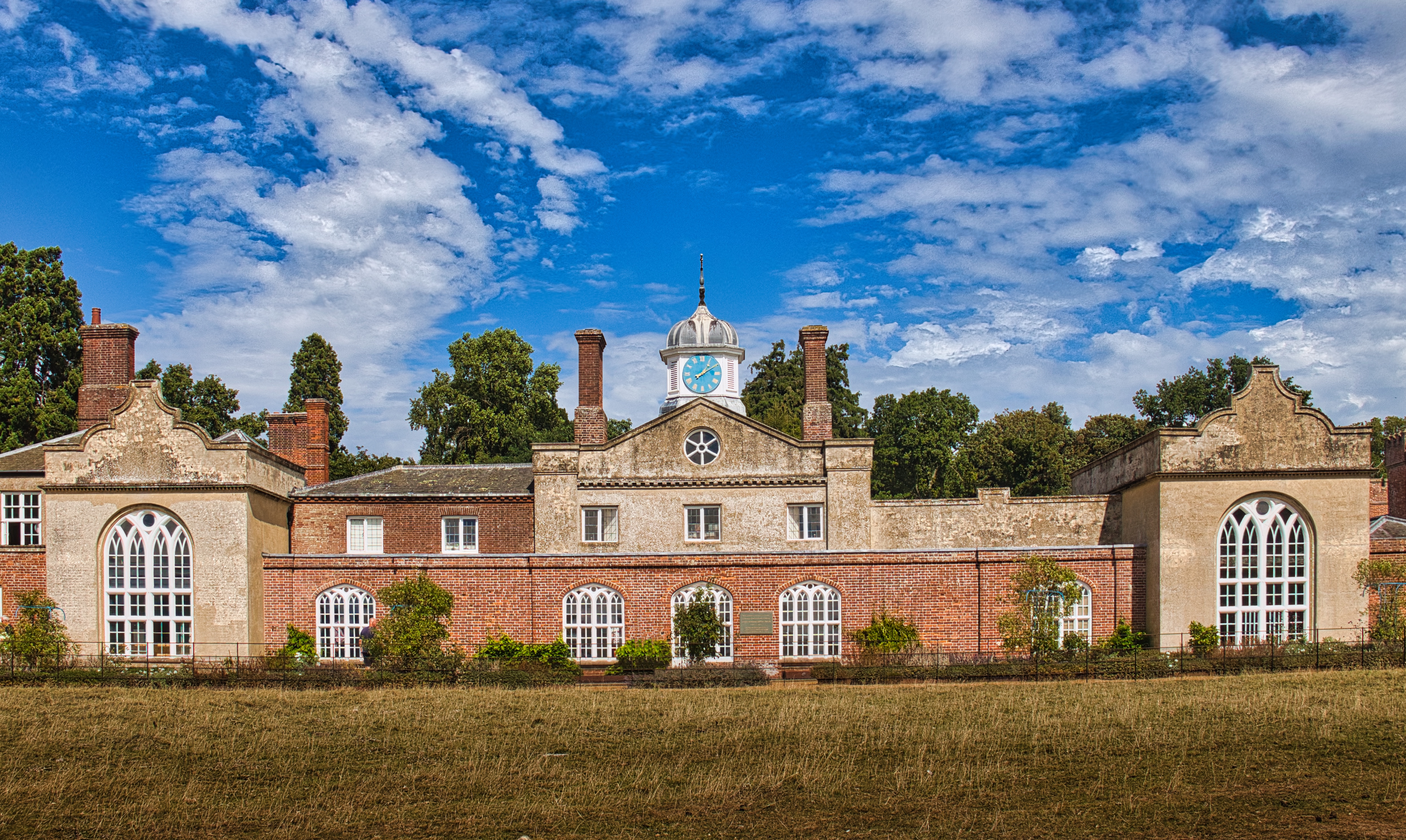 This photograph captures the serene beauty of a historic country estate, bathed in warm golden light of a summer afternoon. The symmetrical composition and rich architectural details evoke a sense of timeless elegance and tranquility. The vibrant blue sky and fluffy white clouds add a touch of serenity, making this image perfect for creating a focal point in a living room or executive office. The warm tones and classic design make it ideal for modern gallery walls, enhancing the aesthetic appeal of any space with its grandeur and charm.