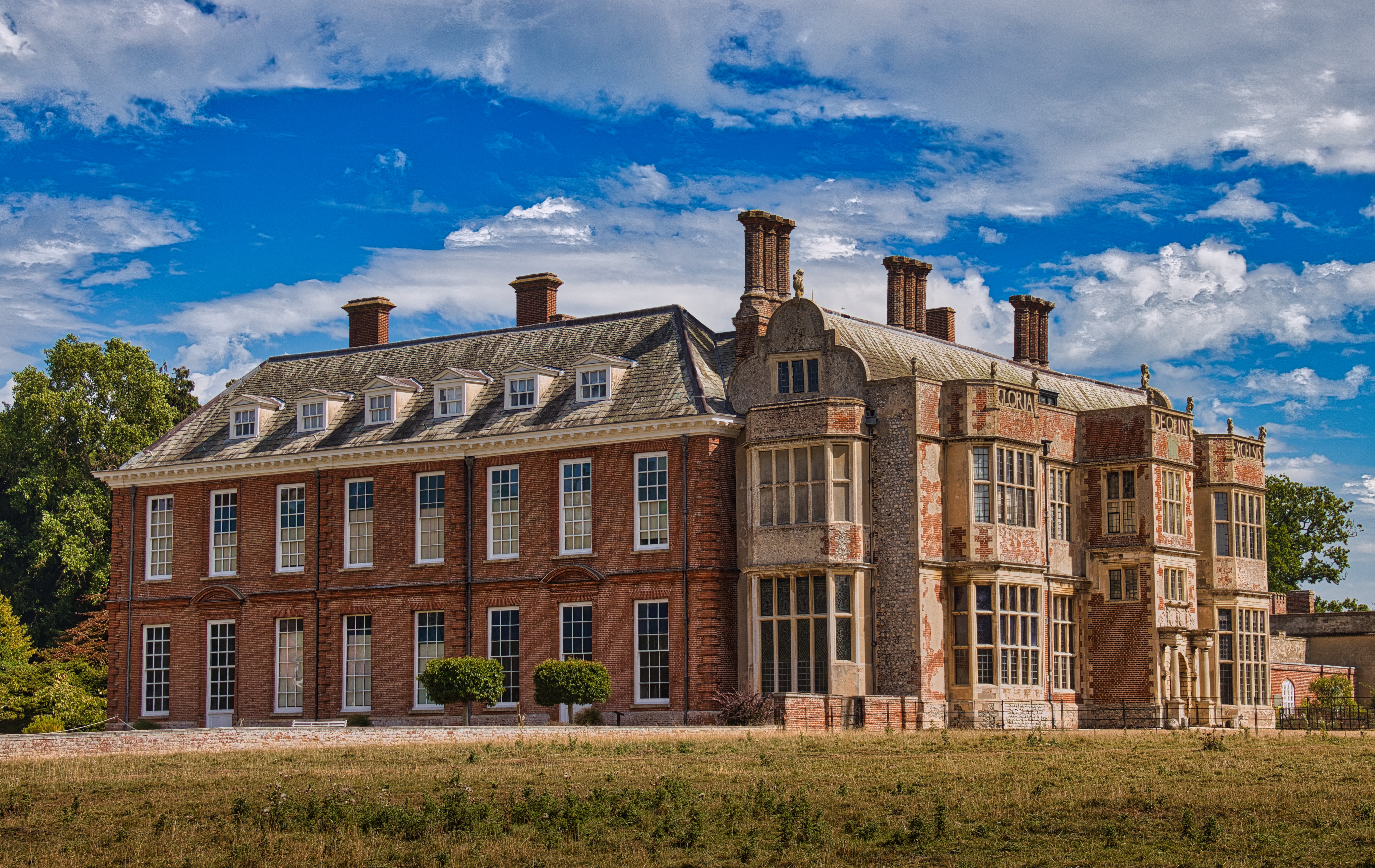 Capture the timeless elegance of this historic manor house, bathed in warm sunlight of a clear blue sky. The rich red brick and intricate architectural details evoke a grandeur of a bygone era, making it a perfect focal point for any sophisticated interior. Display this image in a living room to inspire conversations and admiration, or in an executive office to convey a sense of history and prestige. The serene landscape and vibrant colors make it a versatile piece that enhances any modern gallery wall, adding a touch of classic beauty to your decor.