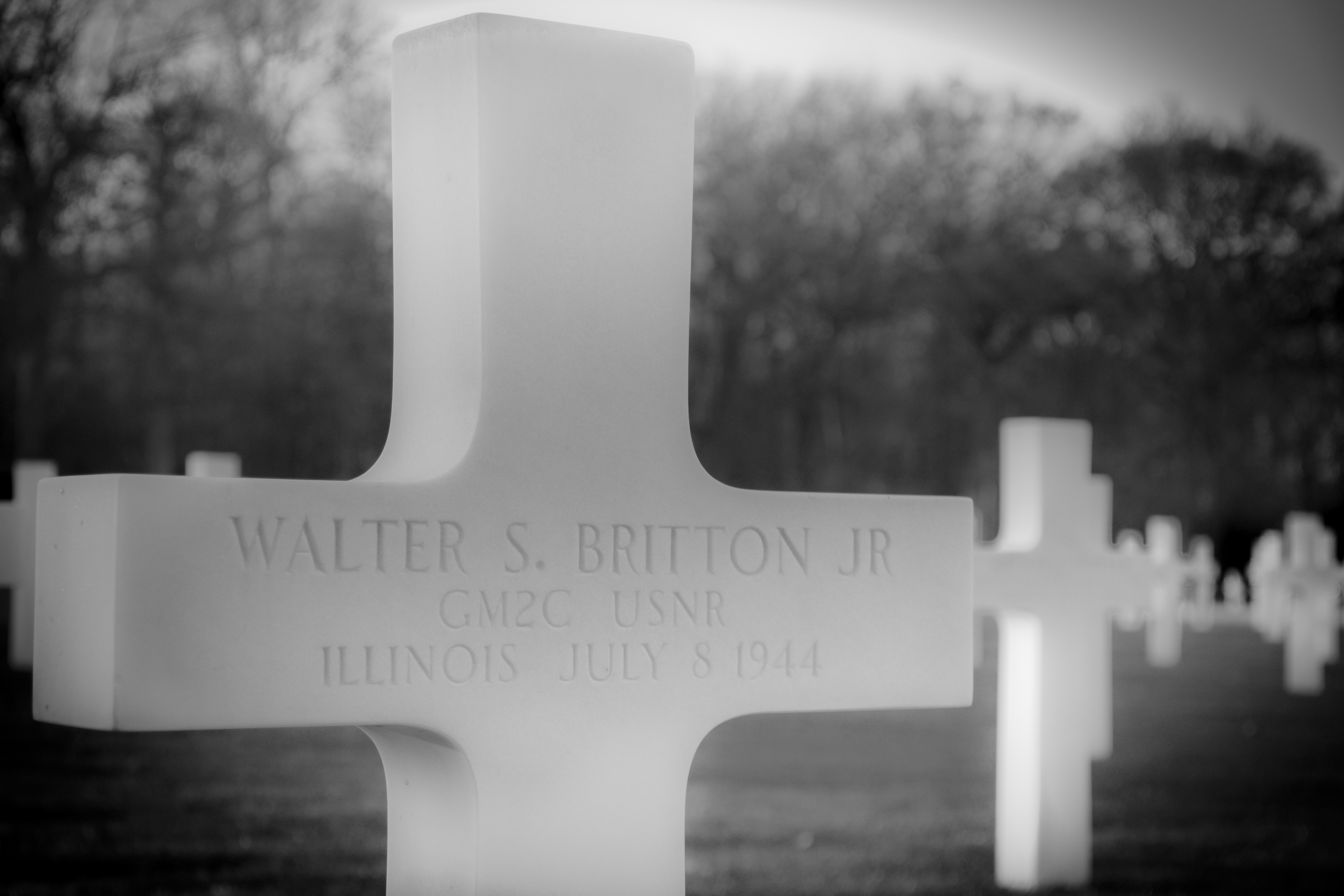 This evocative black and white photograph captures the solemn beauty of a military grave marker, honoring Walter S. Britton Jr. The stark contrast and soft focus create a perfect mood of reverence and remembrance. The composition, with its central cross and blurred background, draws the viewer's eye to the name and service details, evoking a quiet dignity of the fallen. Its aesthetic appeal lies in its timeless quality, making it a ideal piece for a living room, where it can serve as a focal point of reflection and respect. In an executive office, it can symbolize the enduring legacy of service and sacrifice, while in a modern gallery wall, it adds a poignant touch of historical and emotional depth.