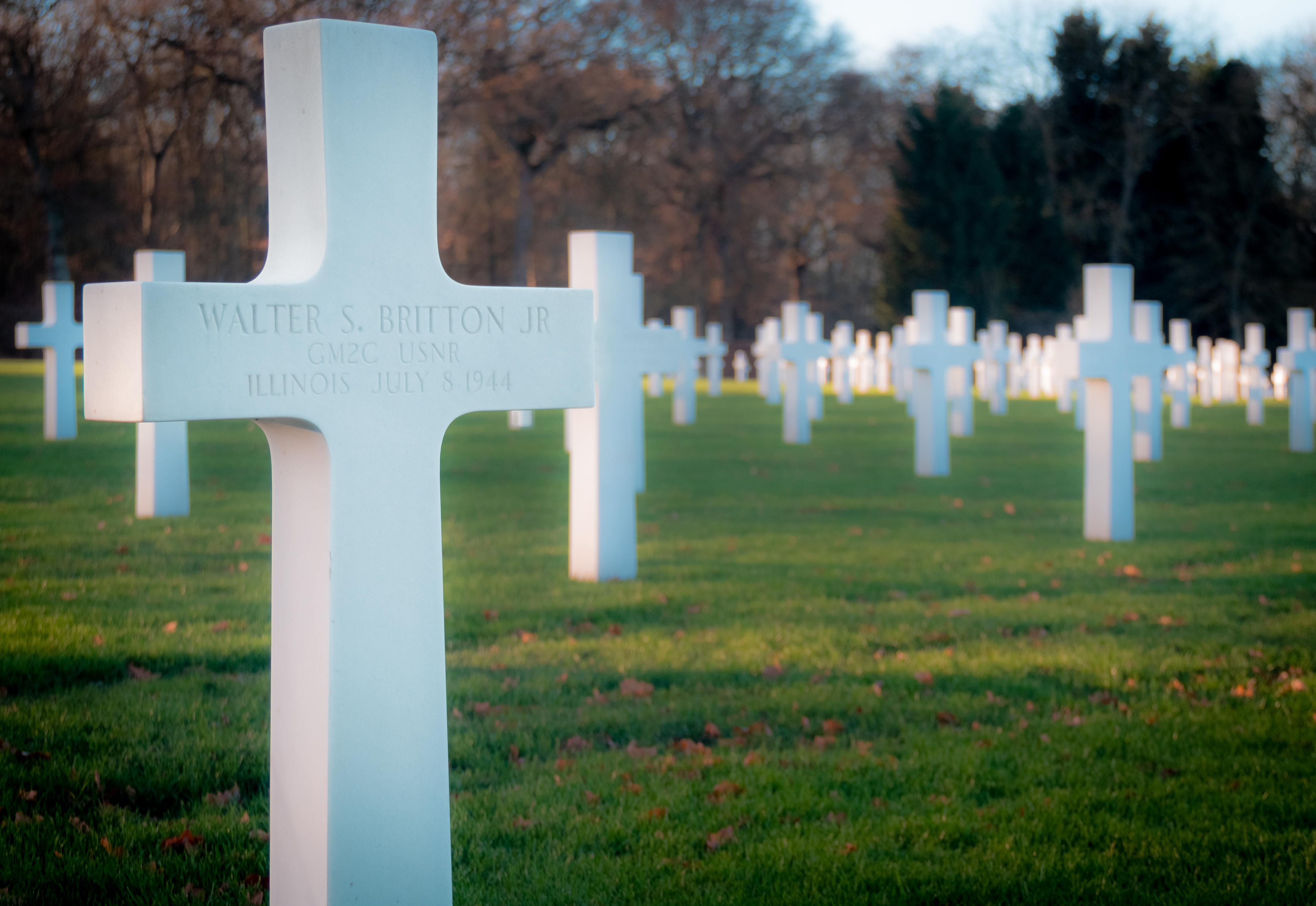 This poignant photograph captures the solemn beauty of a military cemetery, where the soft golden light of the setting sun bathes the white crosses in rest. The composition, with its focus on a cross of Walter S. Britton Jr., evokes a quiet dignity and enduring memory of those who served. The rich, earthy tones of the grass and the bare branches of the trees in background add depth and a sense of timelessness. This image is a perfect piece for any space that honors history and remembrance, whether it be a living room, an executive office, or a modern gallery wall.