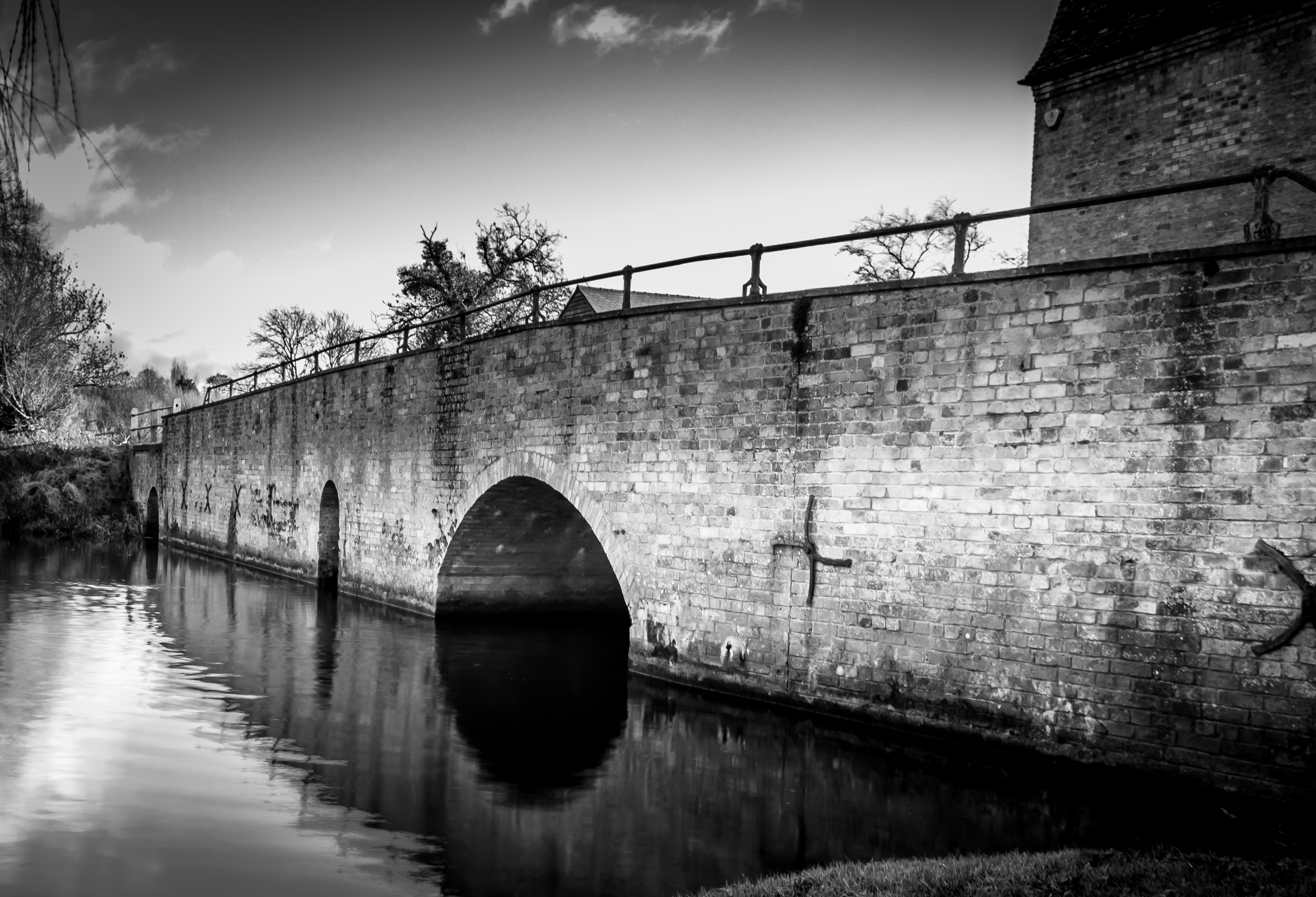 This evocative black and white photograph captures the serene beauty of a historic stone bridge, its arches centerpiece against the tranquil water below. The soft, diffused light enhances the texture of the stone, creating a perfect balance of shadow and light. The composition, with the bridge spanning the frame, draws the viewer's eye across the scene, evoking a feeling of calm and timeless beauty. This image's aesthetic appeal lies in its simplicity and the way it conveys a quiet strength of the structure. Perfect for a living room as a focal piece, or as a executive office's statement wall, it adds a perfect touch of sophistication and historical charm.