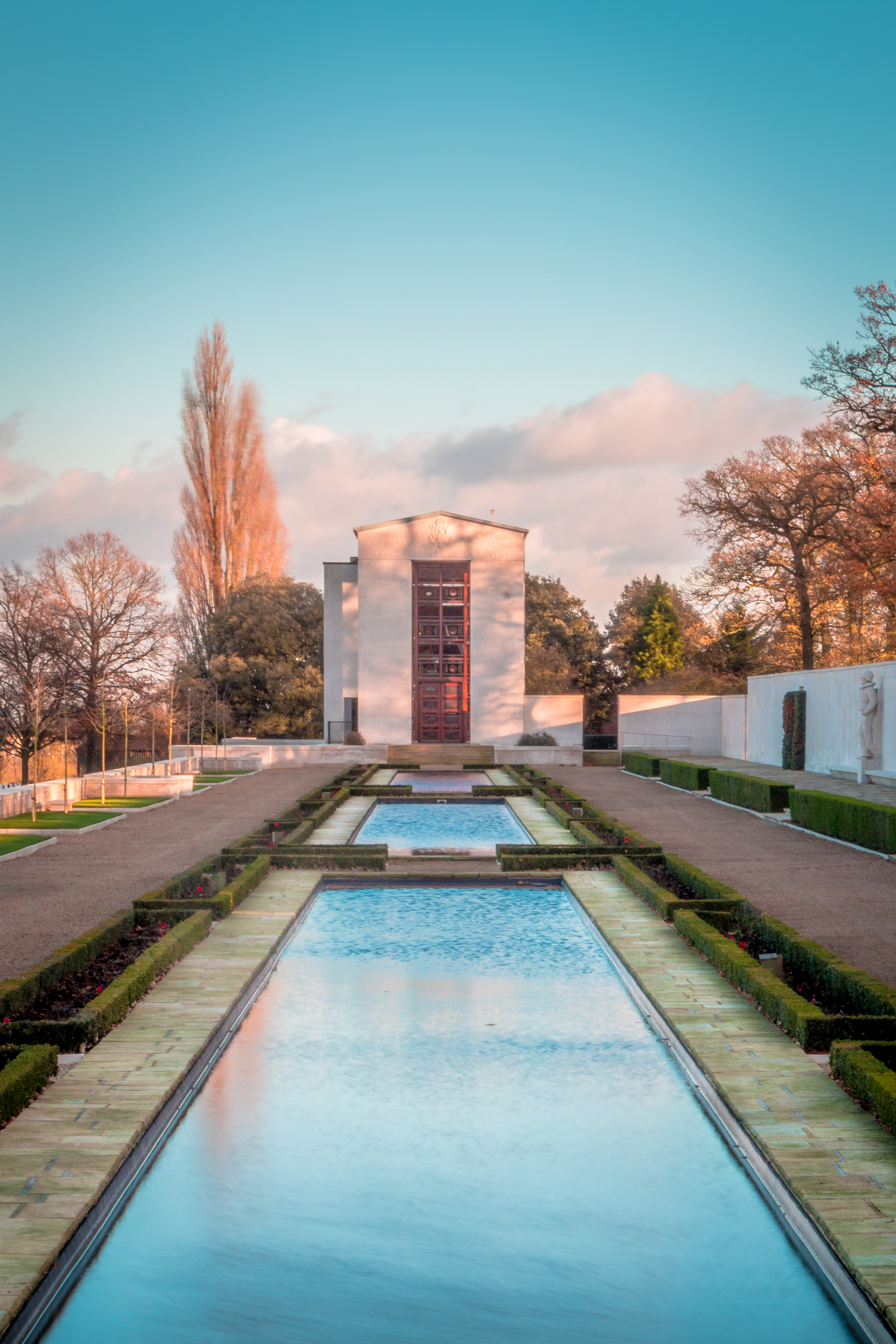 This fine-art print captures the serene beauty of a garden, with its tranquil reflecting pool and the warm glow of the setting sun casting a golden hue over the scene. The composition is balanced, with the architectural structure in focal point, surrounded by lush greenery and a soft, pastel sky. The image evokes a feeling of peace and relaxation, making it an ideal focal piece for a living room or a calming presence in an executive office. The rich, warm tones and the interplay of light and shadow create a visually stunning piece that enhances any aesthetic appeal of any space.