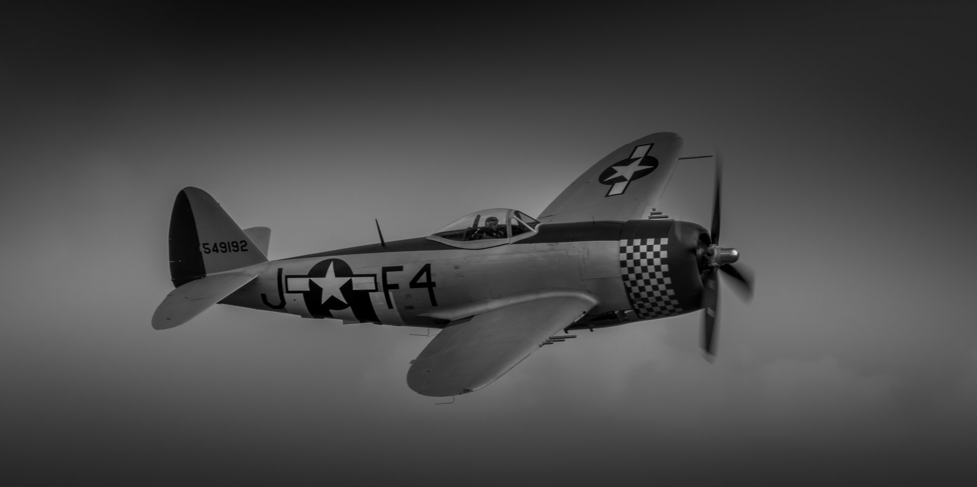 This striking black-and-white photograph captures the essence of a classic World War II-era fighter plane in F4, frozen in mid-flight against a dramatic, moody sky. The composition is balanced, with the aircraft positioned slightly off-center, drawing the viewer's eye across the frame. The lighting, a soft gradient of light and shadow, enhances the texture of the aircraft's surface and the checkerboard pattern on its tail, adding a photograph a timeless appeal of a past. This image evokes a spirit of adventure and the golden age of aviation, making it a perfect piece for any aviation enthusiast or anyone who appreciates the beauty of vintage design. Ideal for a living room focal piece, executive office, or as a centerpiece of a modern gallery wall, this photograph is a perfect blend of history and art.