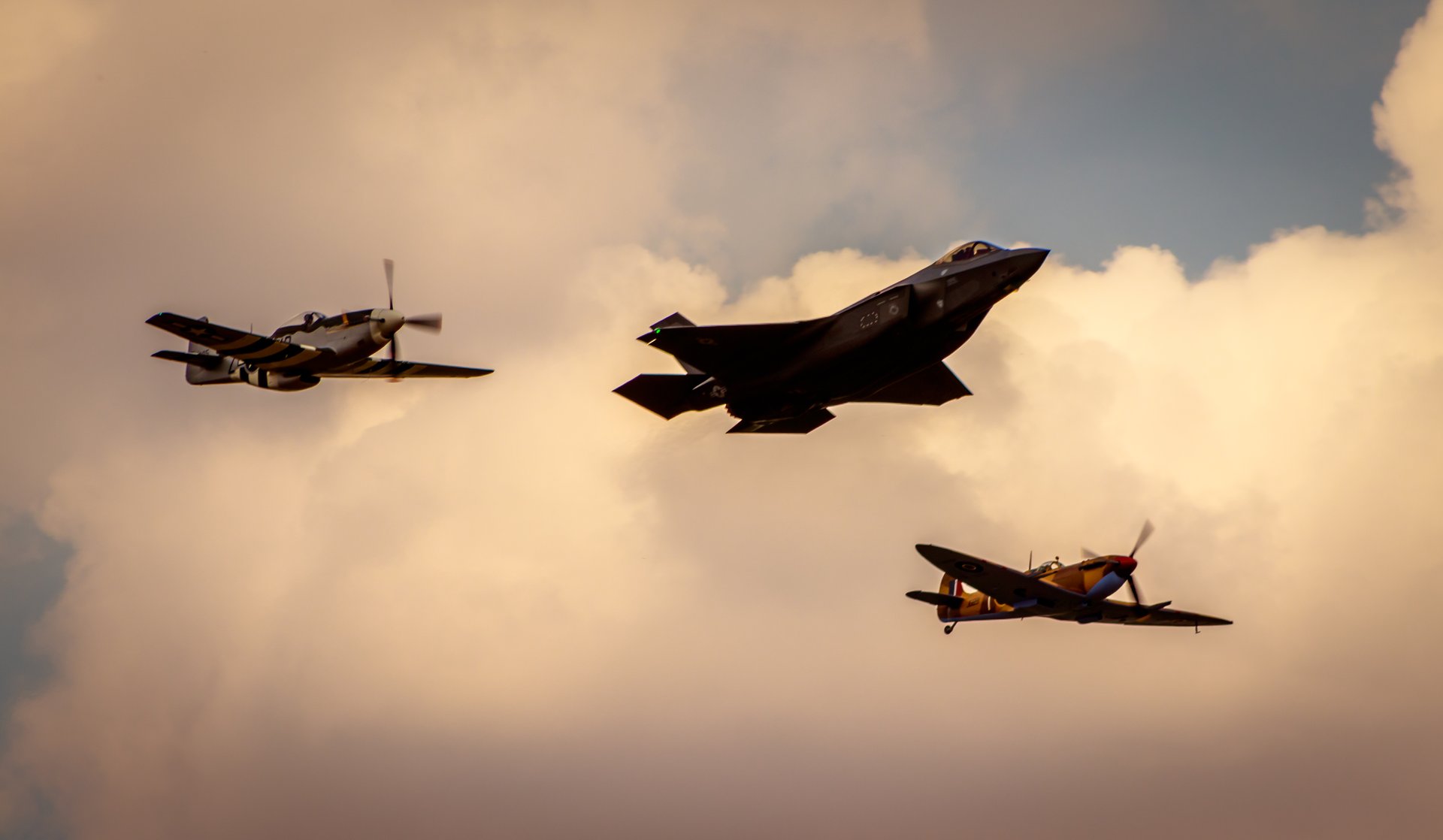 This photograph captures a timeless beauty of aviation history, juxtaposing a sleek, modern fighter jet with vintage World War II-era planes. The warm, golden lighting bathes the scene in, evoking a nostalgia of a bygone era while highlighting the advanced technology of today. The composition, with the planes flying in a triangular formation against a backdrop of a cloudy sky, creates a dynamic and visually striking image. This photograph's aesthetic appeal lies in its ability to bridge the past and present, making it a perfect piece for any aviation enthusiast or anyone who appreciates the art of flight. Ideal for placement in a living room as a focal piece, or in executive office to inspire innovation and admiration for aviation's evolution.