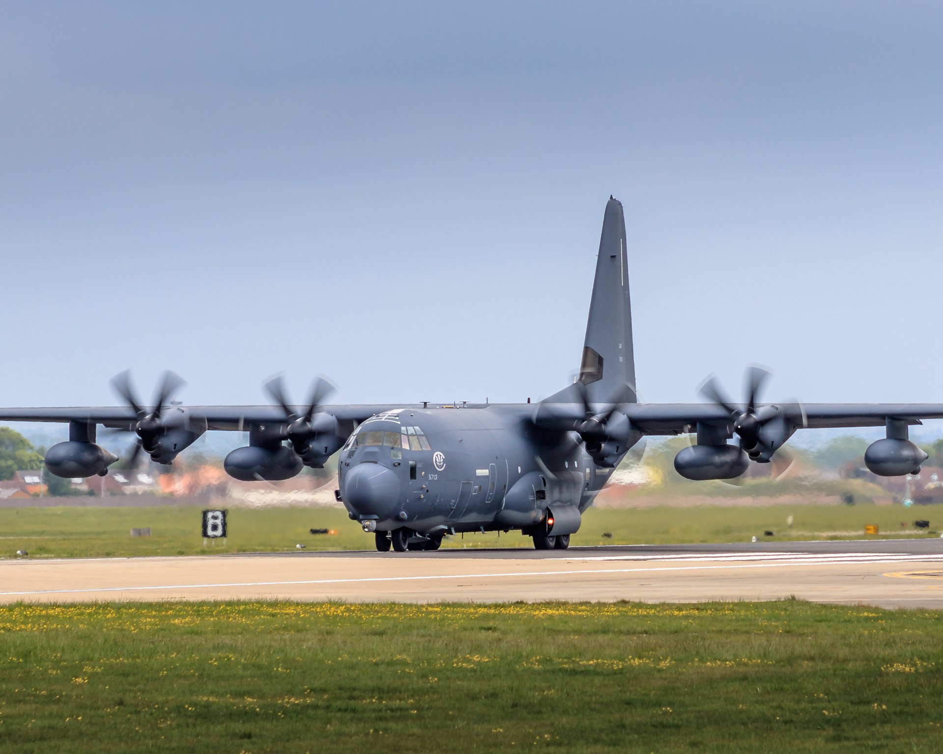 This photograph captures the raw power and precision of a C-130 Hercules in motion, showcasing its robust design and the dynamic energy of its four powerful engines. The image is bathed in natural light, highlighting the aircraft's sleek, military gray paint and the intricate details of its structure. The composition, with the aircraft positioned centrally, draws the viewer's eye across the frame, emphasizing its imposing presence. This background of a runway and distant landscape adds depth and context, making this image not only a centerpiece of a modern gallery wall but also focal point of a executive office, symbolizing strength and innovation.