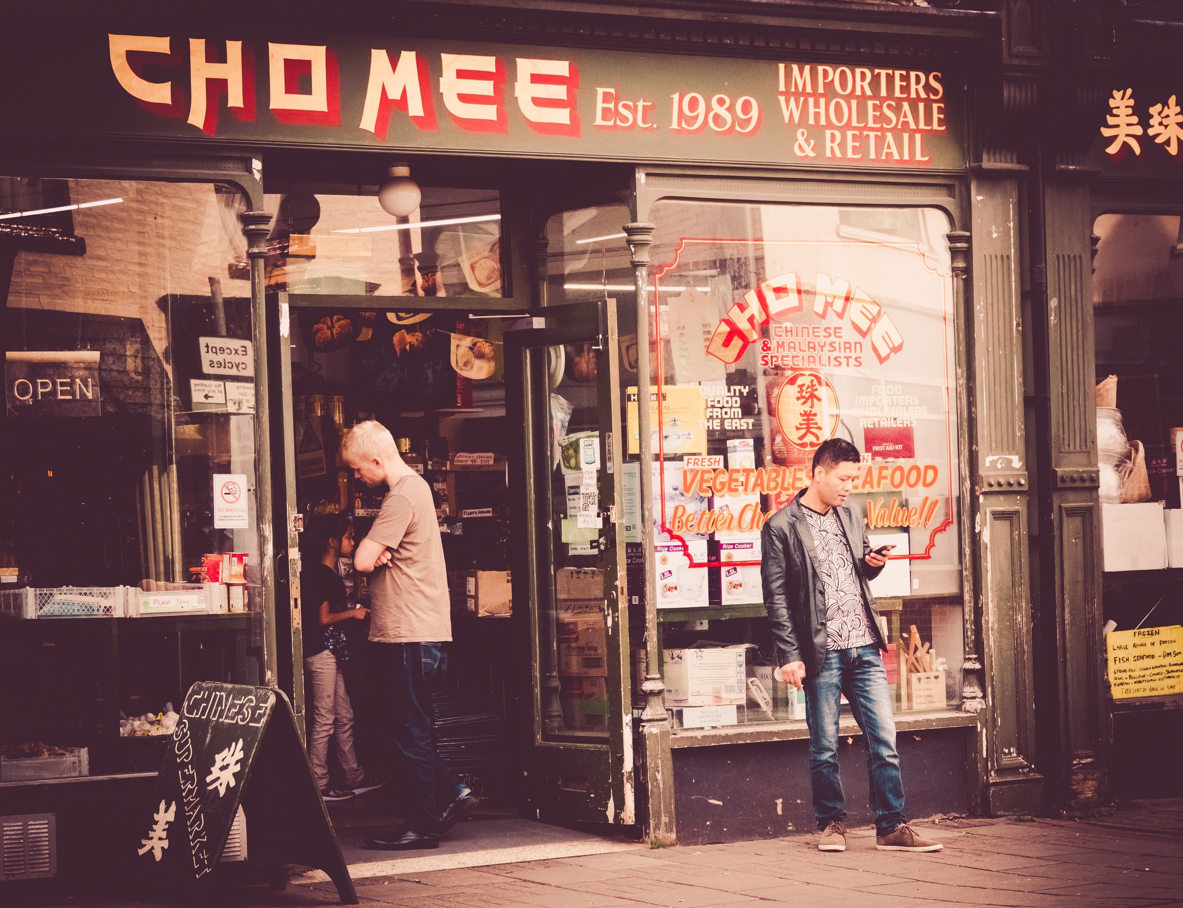 This photograph captures the timeless charm of a 'Chomee' store, established in 1989, with its inviting storefront and bustling street life. The warm, golden lighting enhances the nostalgic feel, while the composition draws the viewer's eye through the glass doors and into the inviting interior. The aesthetic appeal lies in its retro vibe, perfect for adding character of a living room or as a focal piece in executive office. The image evokes a essence of a past, making it a perfect addition to a modern gallery wall, where it can inspire a soul and spark conversations.