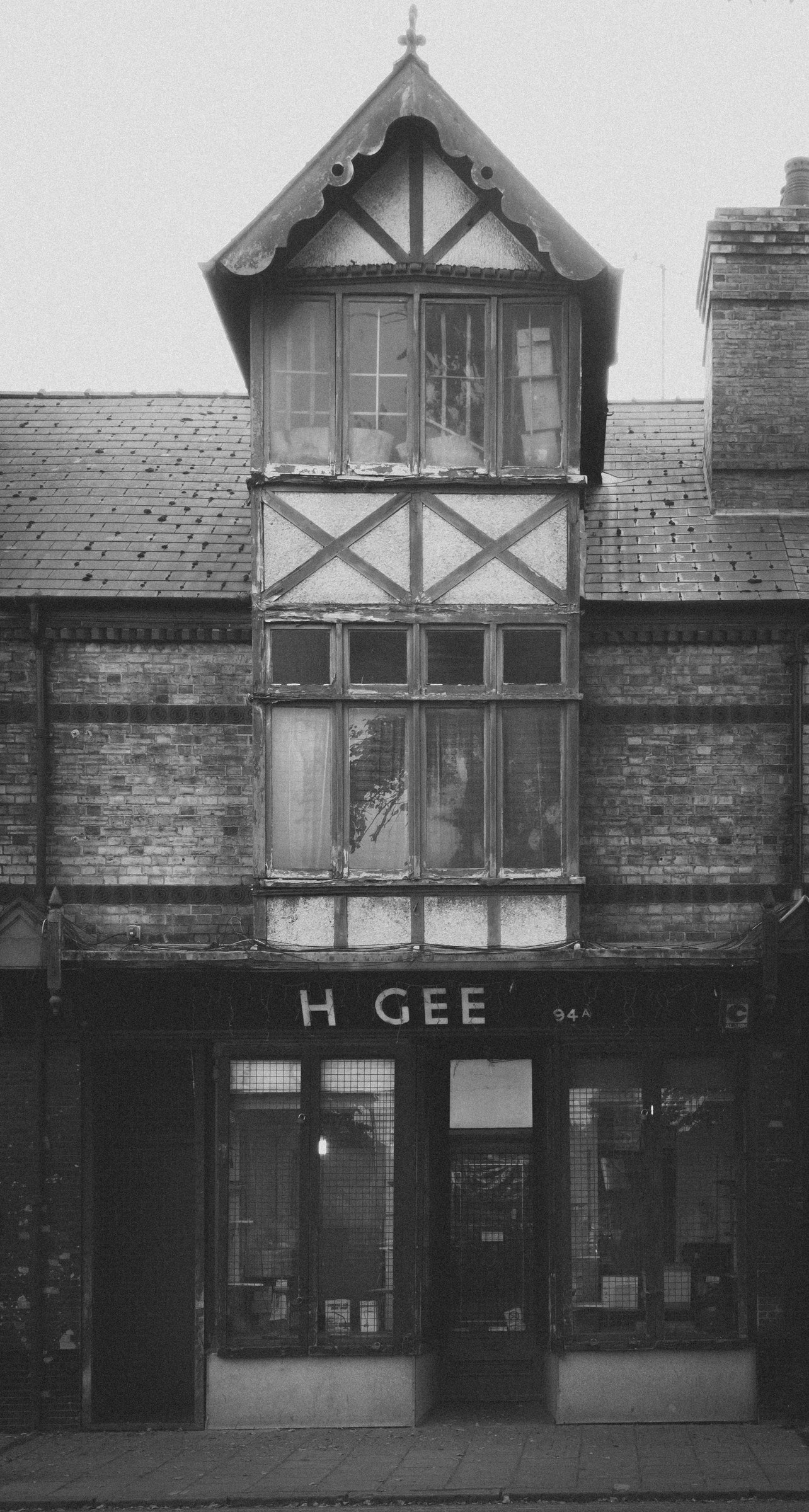 This evocative black and white photograph captures the timeless charm of a Victorian-era building, its architectural details and intricate woodwork standing out against the stark contrast of the monochrome palette. The composition, with its symmetrical facade and prominent gabled window, invites the viewer to explore the rich history and craftsmanship of the era. The soft, diffused lighting enhances the texture of the brick and wood, creating a perfect backdrop for a bold, modern lettering of 'H GEE'. This image's aesthetic appeal lies in its ability to evoke a past while resonating with contemporary tastes, making it an ideal focal piece for a living room or a executive office, where it can inspire a sense of history and sophistication.