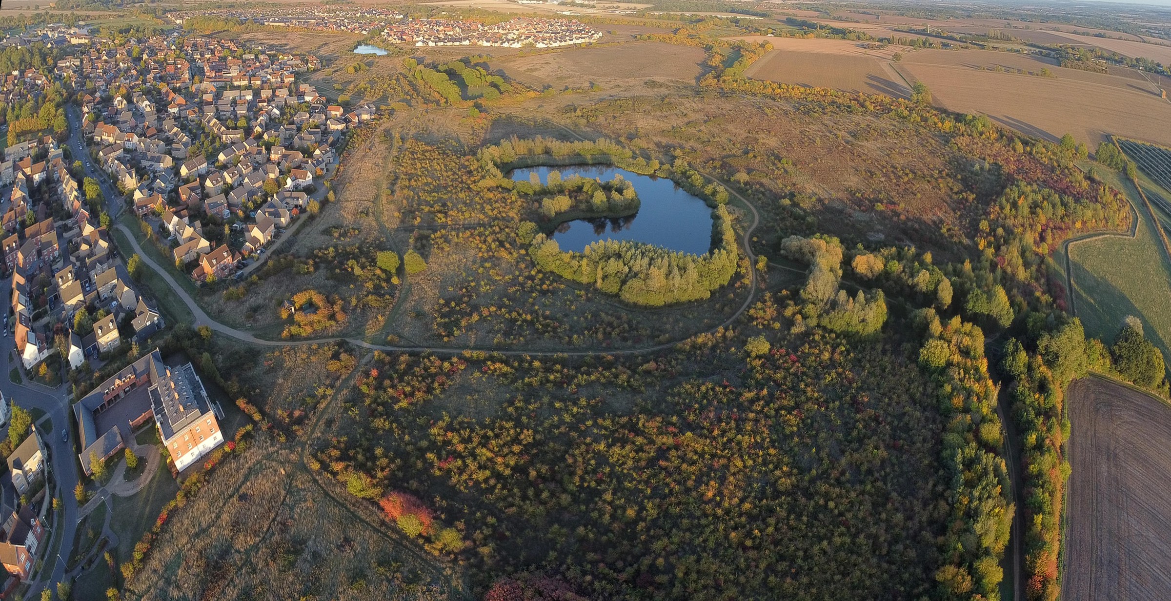 This breathtaking aerial photograph captures the serene beauty of a transition between urban and natural landscapes, bathed in the warm, golden hues of the setting sun. The composition beautifully juxtaposes the structured, geometric lines of the town with the organic, flowing contours of the surrounding fields and forested areas. The rich autumnal colors of the foliage add a perfect touch of vibrancy and warmth, making this image a perfect focal point for any modern gallery wall or a executive office, where it can inspire a spirit of balance and harmony between nature and human habitation.
