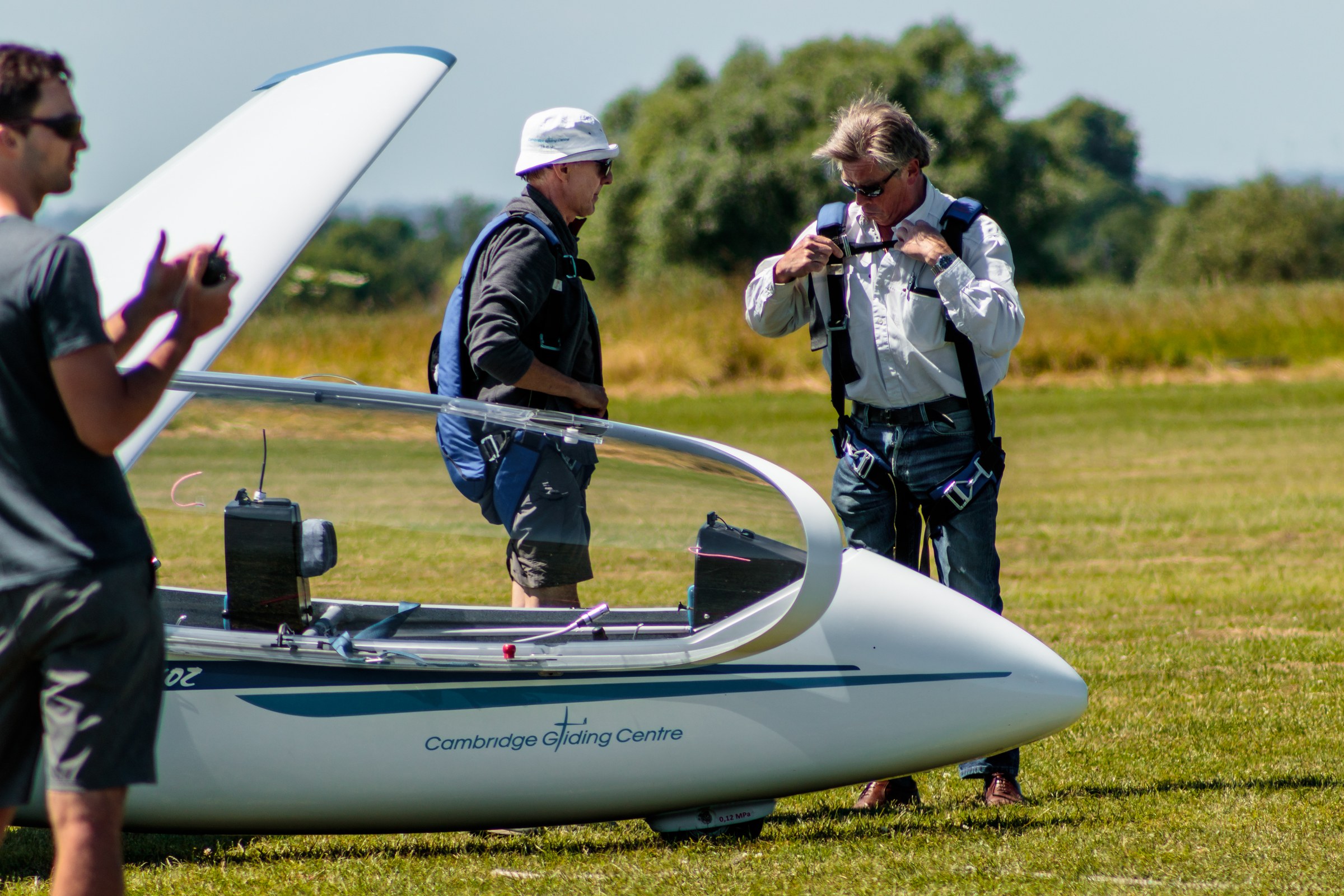 This photograph captures the tranquil essence of a gliding adventure, with a soft, natural light of a sunny day casting a scene in in a perfect balance of light and shadow. The composition centers on the sleek, white glider, its pointed nose and streamlined design drawing the eye, while the figures in background add a human element, suggesting preparation and anticipation. The vibrant green field and the distant trees provide a serene backdrop, evoking a feeling of freedom and escape. This image is ideal for a living room focal piece, where it can inspire a spirit of adventure and tranquility, or for an executive office, where it can serve as a perfect reminder of the calm and focus required in leadership.
