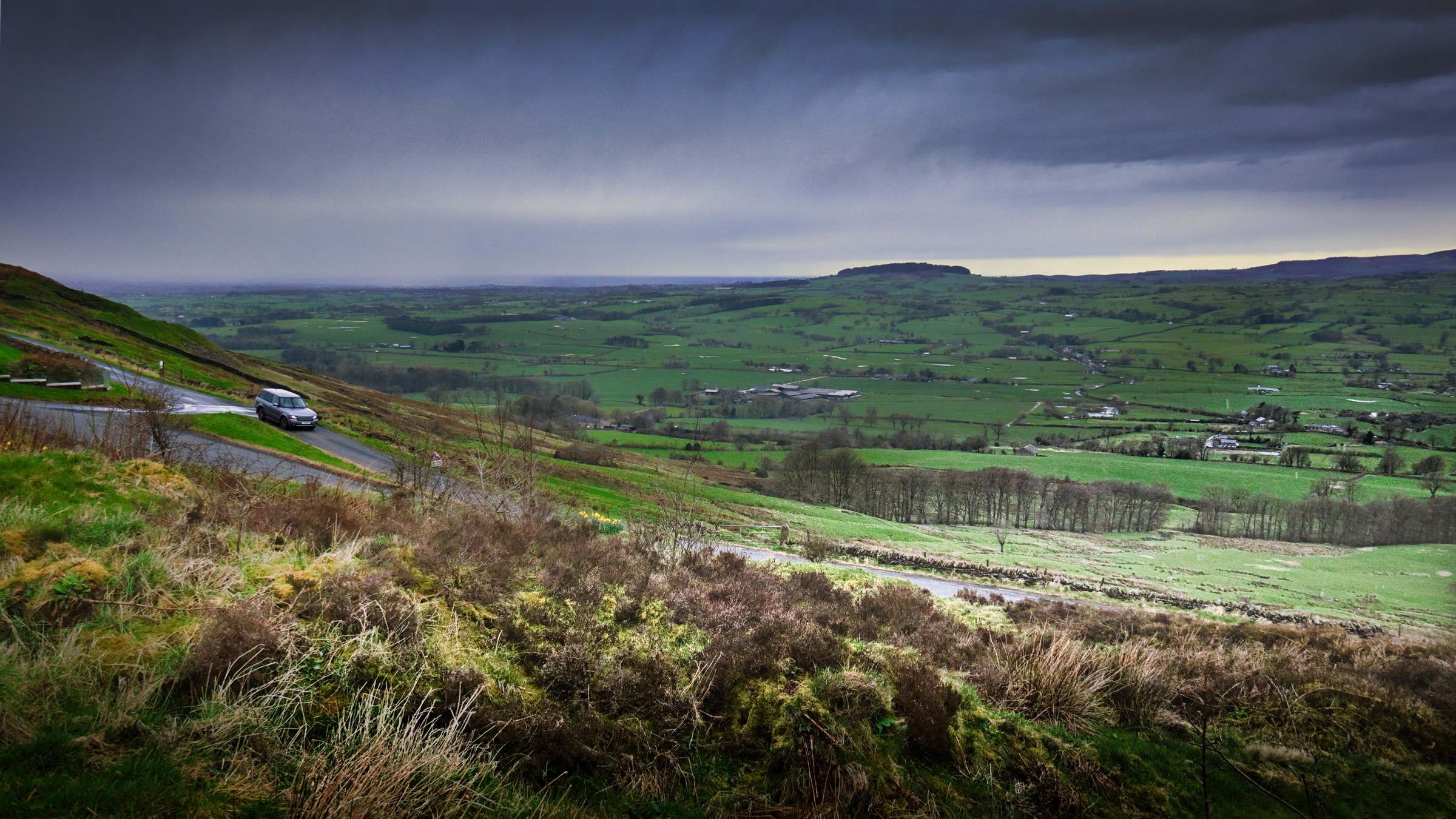 This photograph captures a serene valley under a dramatic, stormy sky, evoking a sense of tranquility and awe. The composition beautifully balances the foreground's rugged, grassy hillside with the expansive, lush valley in the background. The interplay of light and shadow, with the dark clouds casting a moody ambiance, adds depth and drama to the scene. The aesthetic appeal lies in its natural beauty and the subtle contrast between the earthy tones and the vibrant greens, making it a perfect piece for a living room as a focal point or for the executive office to inspire calmness and creativity.