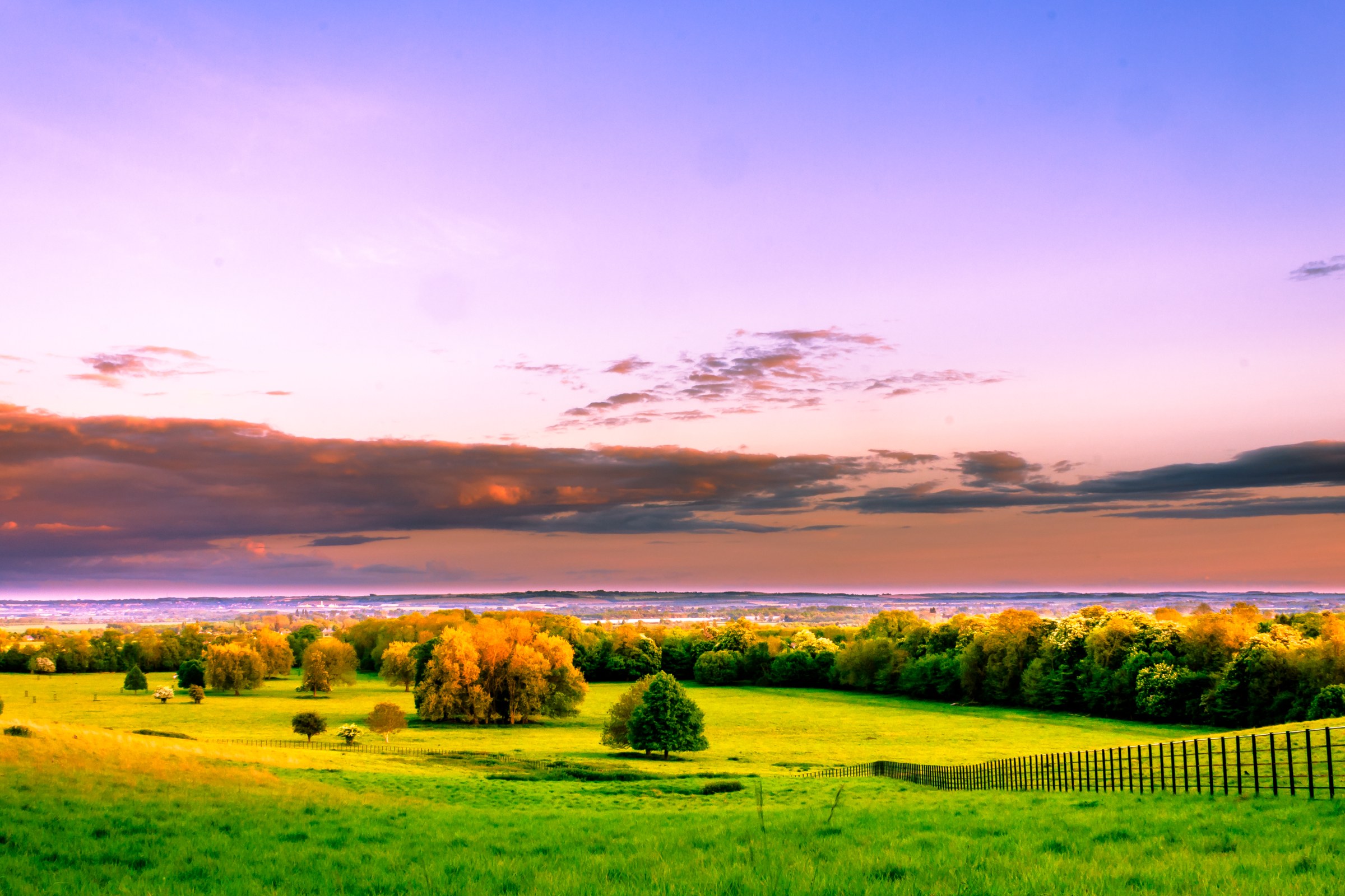 This breathtaking photograph captures the tranquil beauty of a pastoral sunset, where the sky transitions from a warm hues of orange and pink to the cool tones of purple and blue. The composition is balanced with a gentle curve of a fence leading the eye towards the horizon, framed by lush green fields and a silhouettes of trees. The vibrant colors and serene atmosphere make it an ideal piece for enhancing any living room or executive office, serving as a focal point that evokes a calmness and beauty of nature. Perfect for those who appreciate the art of landscape photography and the calming effect of natural scenery.