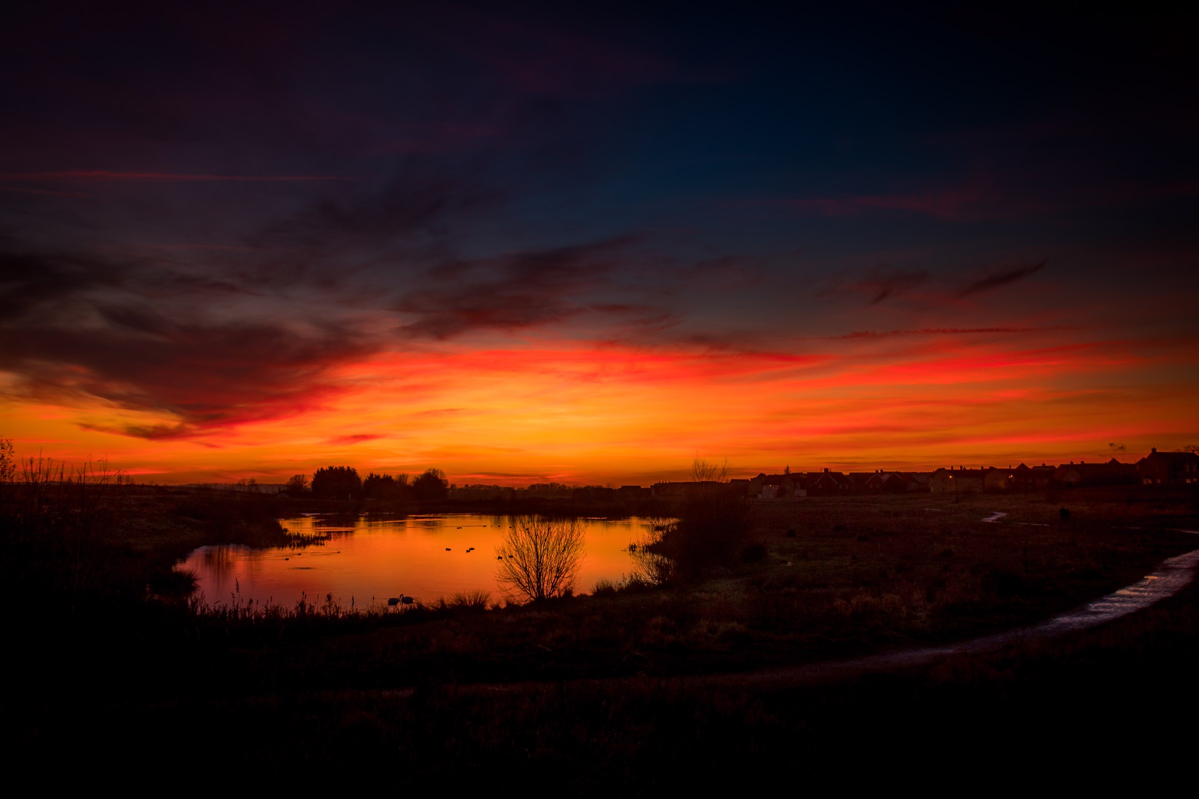 This breathtaking photograph captures the mesmerizing beauty of a sunset, where the sky is ablaze with fiery hues of orange, red, and purple, reflected in a tranquil waters below. The composition is balanced with a silhouette of a landscape on the left, creating a perfect harmony of light and shadow. The rich, warm tones of the sunset evoke a sense of peace and tranquility, making it an ideal piece for any living room or office space. Display it as a focal point in your living room to enhance the ambiance or in your executive office to inspire calm and focus.