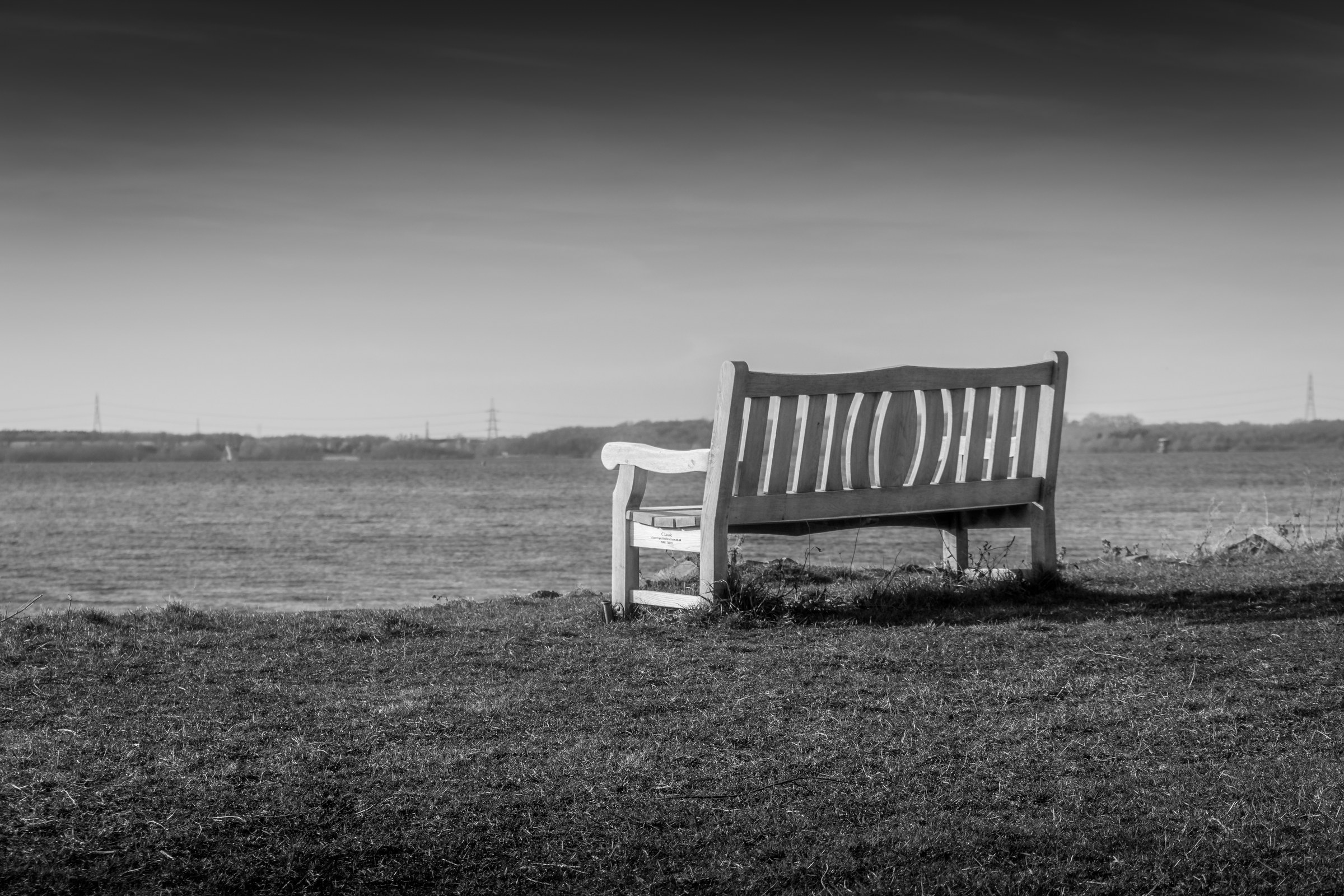 This evocative black and white photograph captures the serene solitude of a lone bench in an open field, evoking a quiet contemplation of nature. The composition, with the bench slightly off-center, draws the viewer's eye into the vast, open landscape, creating a perfect balance between human presence and the grandeur of the natural world. The soft, diffused lighting enhances the timeless quality of the scene, making it a ideal piece for a living room, where it can serve as a focal point of a space, or for the executive office, where it can inspire moments of reflection and calm amidst the hustle and bustle.
