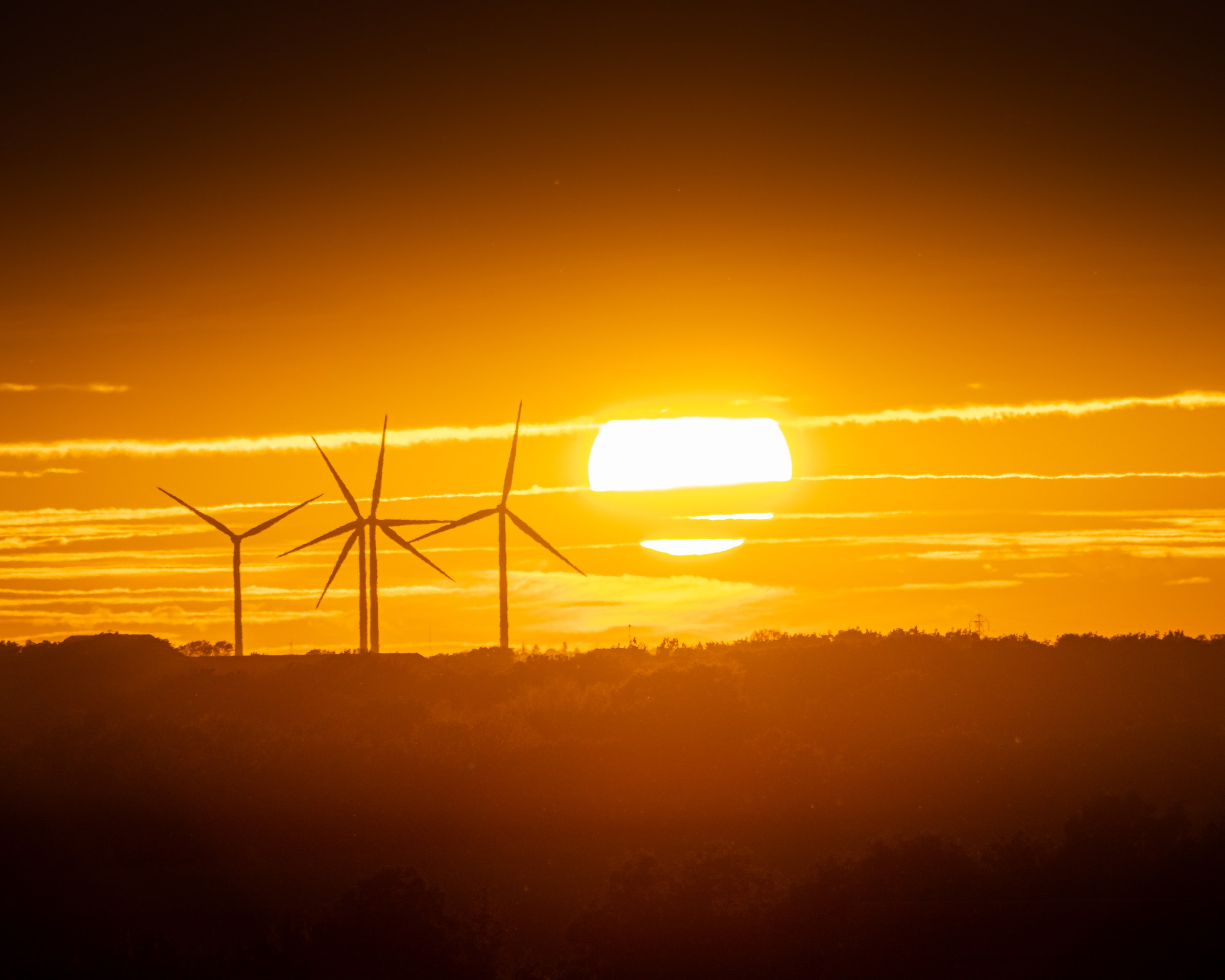 A sunset scene featuring wind turbines silhouetted against a vibrant orange and yellow sky, with the sun partially hidden behind the horizon.