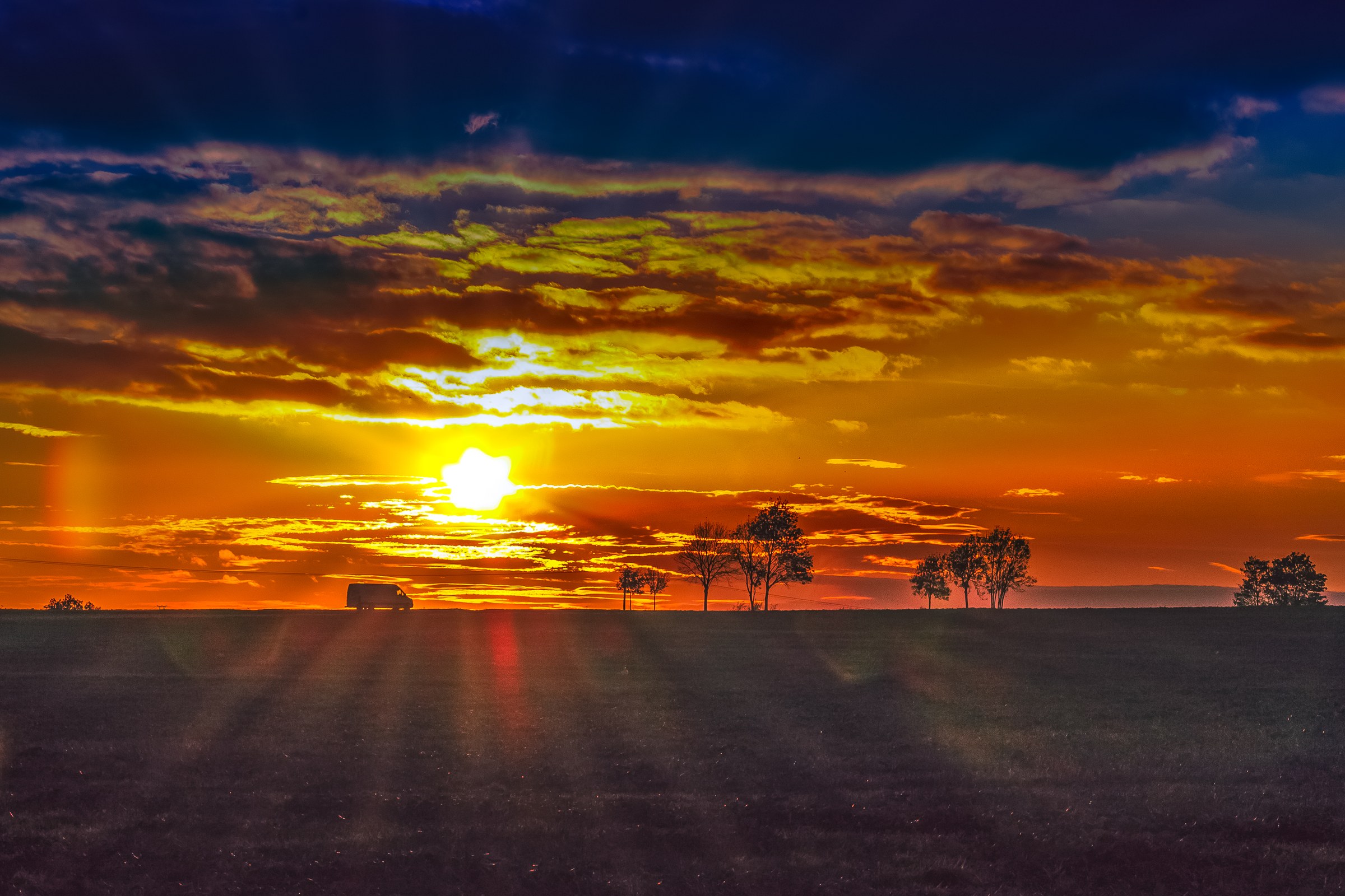 A serene sunset scene with a lone tree and a distant vehicle silhouetted against a vibrant sky, creating a perfect balance of light and shadow.