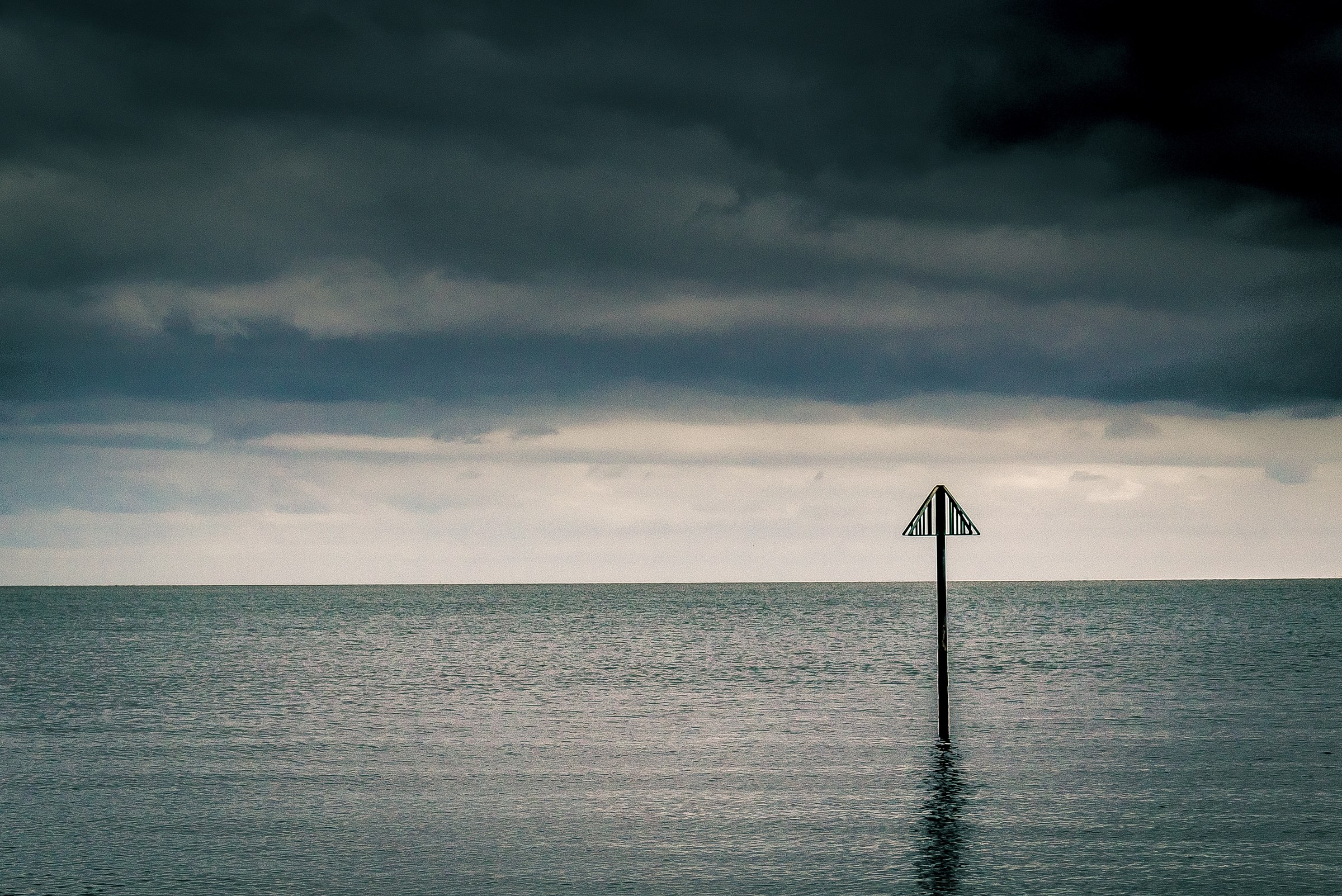 A solitary marker stands in calm waters under a dramatic, overcast sky, creating a contemplative and serene scene.