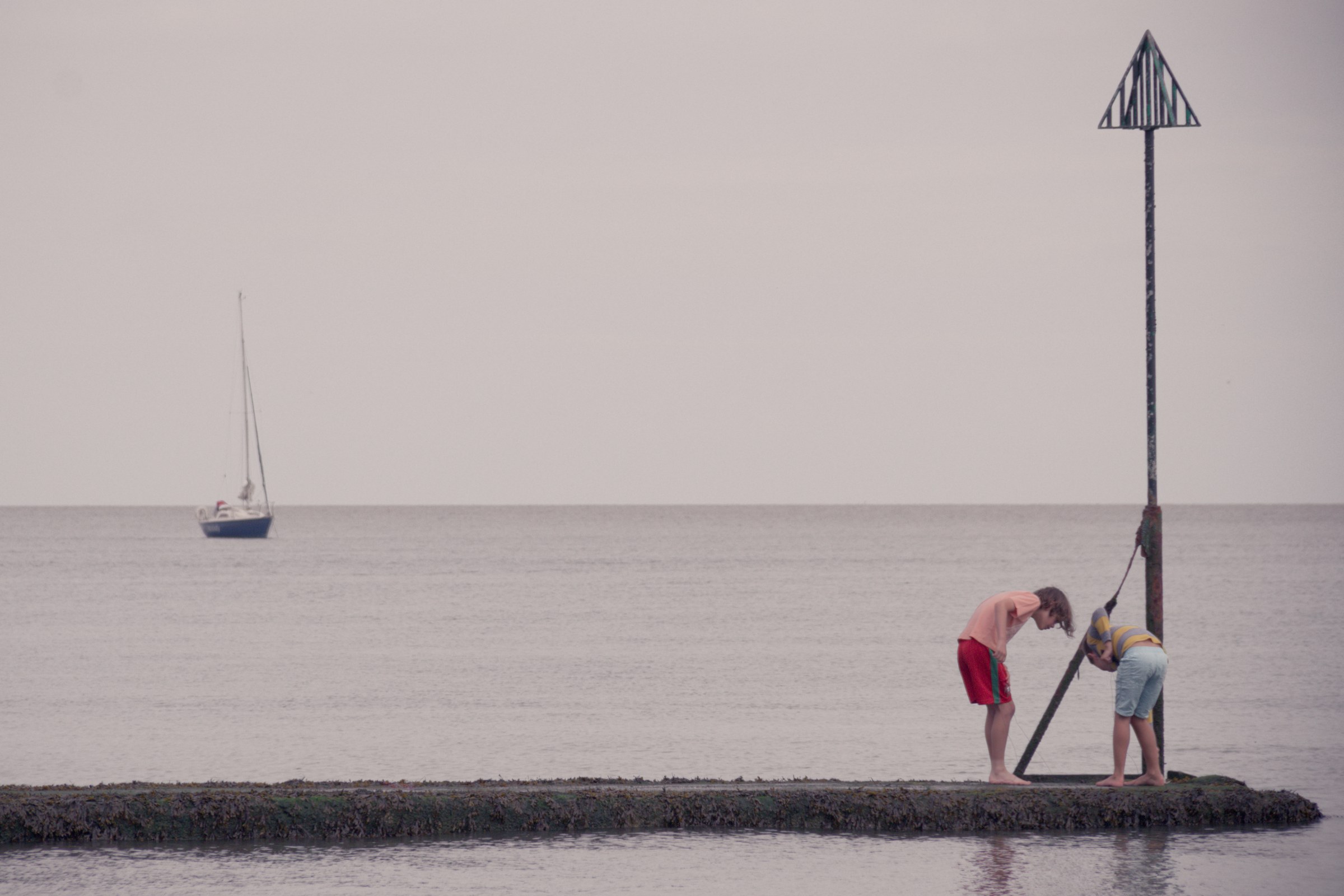 This evocative photograph captures a tranquil coastal scene, where two individuals are engaged in a quiet moment on a small pier. The soft, diffused light of an overcast sky enhances the calm and reflective mood, while the vast expanse of the sea and the distant sailboat add a sense of solitude and vastness. The composition, with its balanced elements and leading lines, draws the viewer's eye from the foreground to the horizon, creating a perfect balance of human interaction and natural beauty. This aesthetic appeal lies in its simplicity and the timeless quality it evokes, making it a perfect piece for a living room or a executive office, where it can serve as a focal point, inspiring a calm and reflective spirit it portrays.