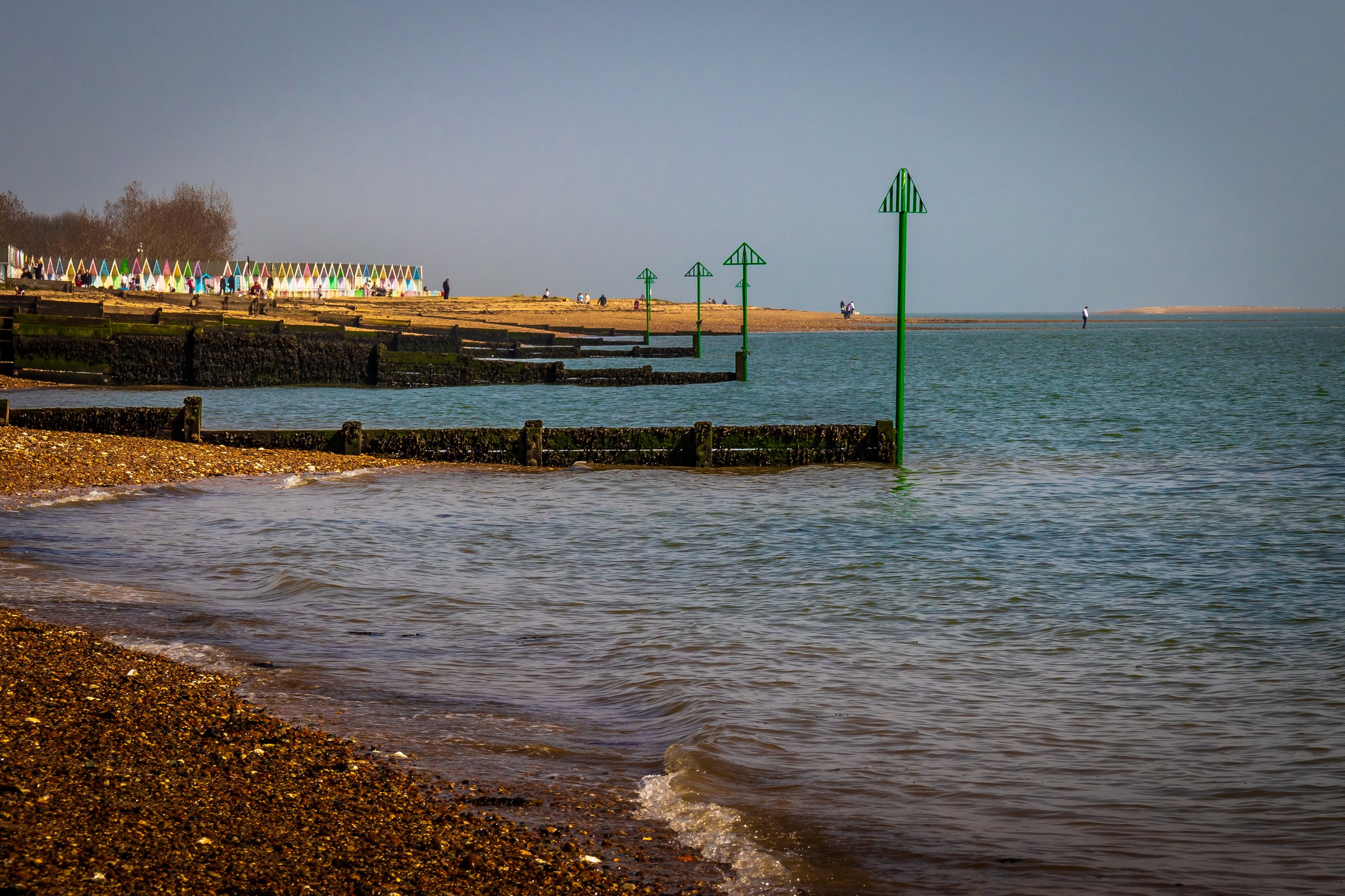 This fine-art print captures the tranquil beauty of a coastal scene, where gentle waves lap against a pebble beach under a soft, diffused light of a clear day. The composition is balanced, with the vibrant green posts of the pier leading the eye towards the horizon, creating a perfect focal point. The muted colors and soft lighting evoke a calm and peaceful mood of a seaside, making it an ideal piece for any living room or executive office. Hang it as a centerpiece of a wall to instantly transform the space into a a serene retreat.