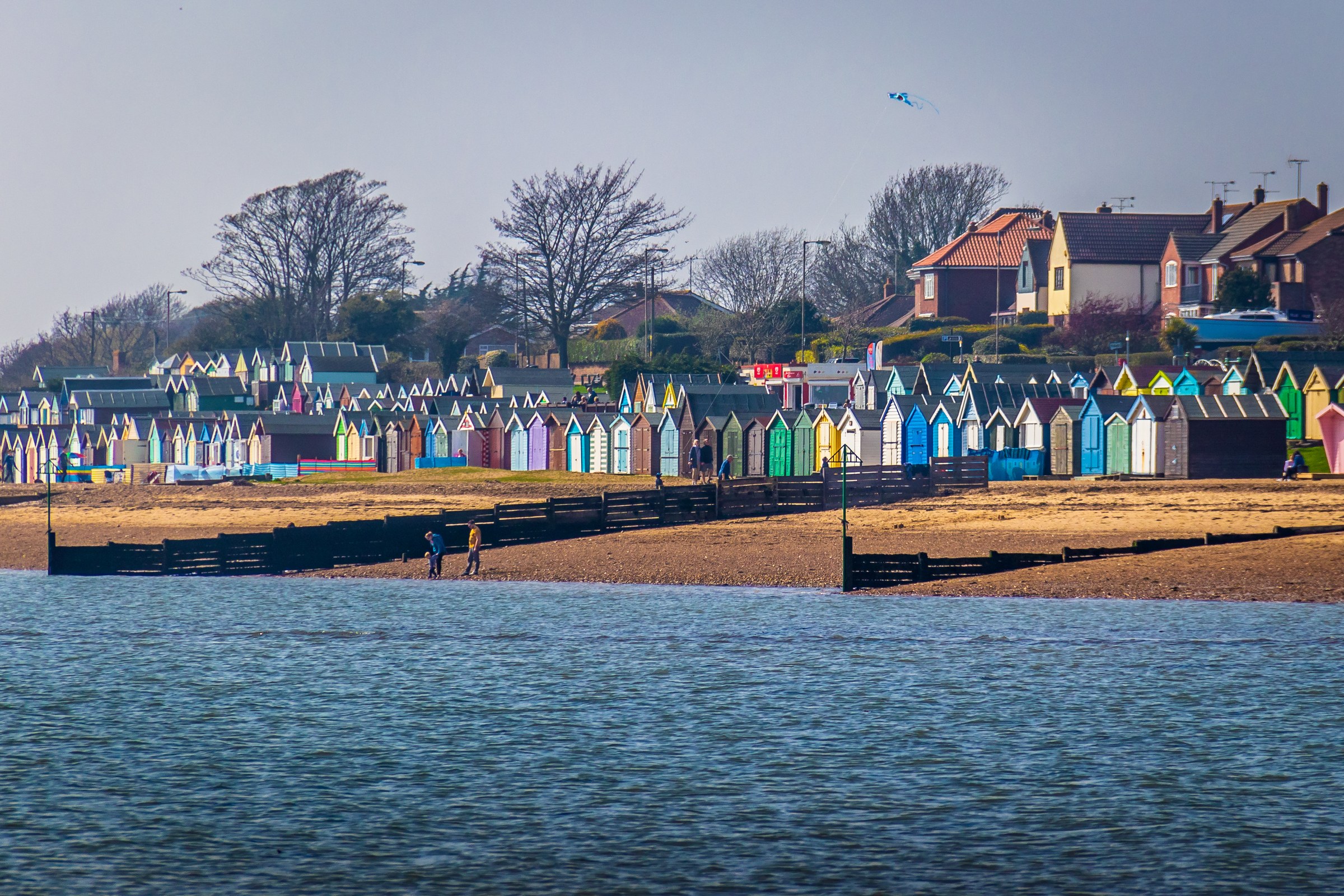 Capture the essence of a tranquil coastal retreat with this vibrant photograph of colorful beach huts lining a shore. The warm, natural light bathes the scene in, highlighting the rich hues of the huts and the serene blue of the water. The composition, with its balanced horizon and leading lines, draws the viewer into the peaceful atmosphere. Perfect for a living room as a focal piece, evoking a feeling of a beach in comfort of home.