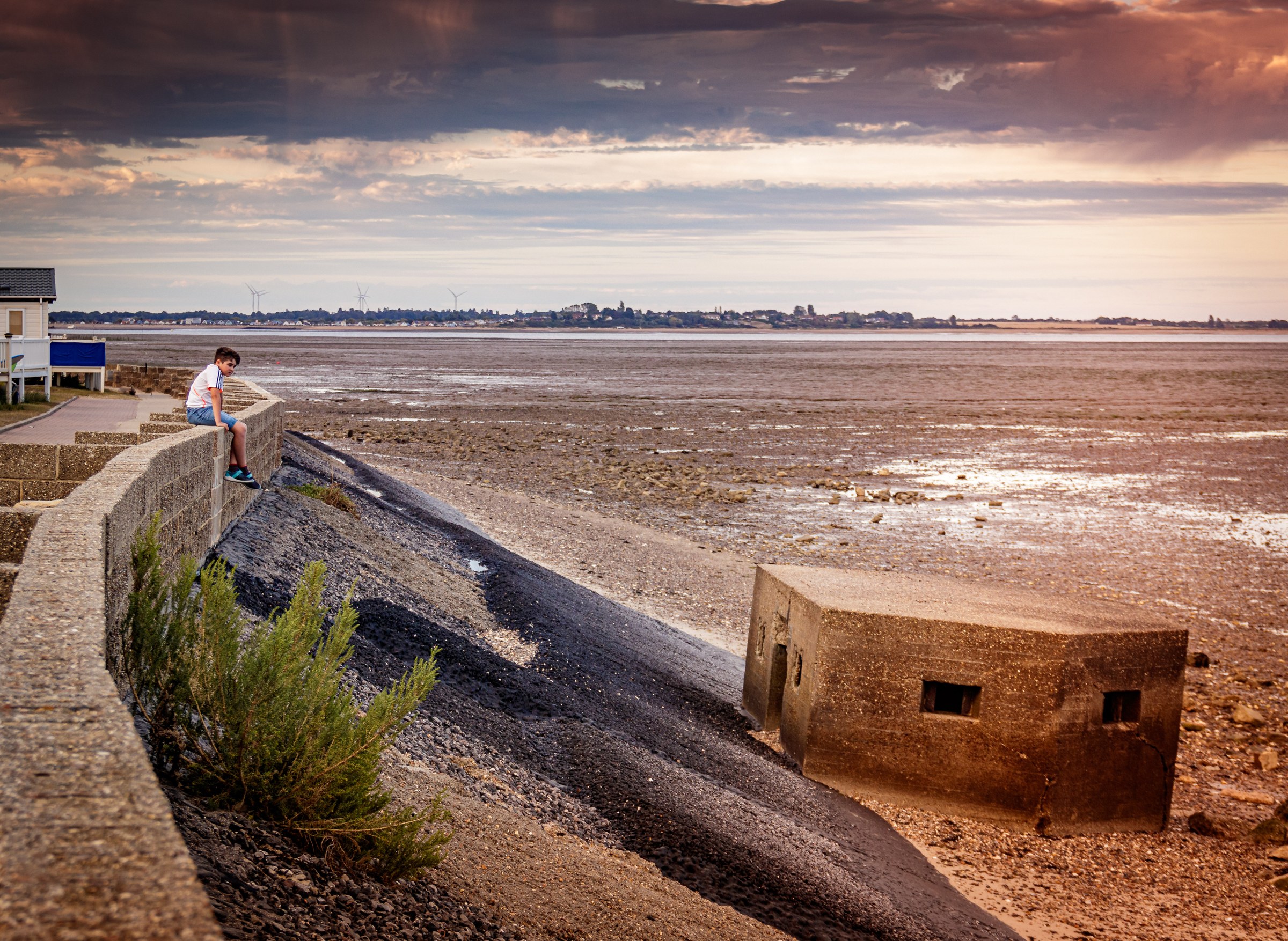 This evocative fine-art photograph captures the tranquil beauty of a seaside at low tide, where the vast expanse of the wetlands meets the horizon under a soft, diffused light of a setting sun. The composition is masterfully balanced, with a solitary figure on the left adding a human element, while the rugged concrete structure on the right provides a stark contrast to the natural landscape. The rich, warm tones and the interplay of light and shadow create a perfect mood of peaceful solitude. This image is ideal for wall decor in living room, creating a perfect focal point that invites contemplation and relaxation.
