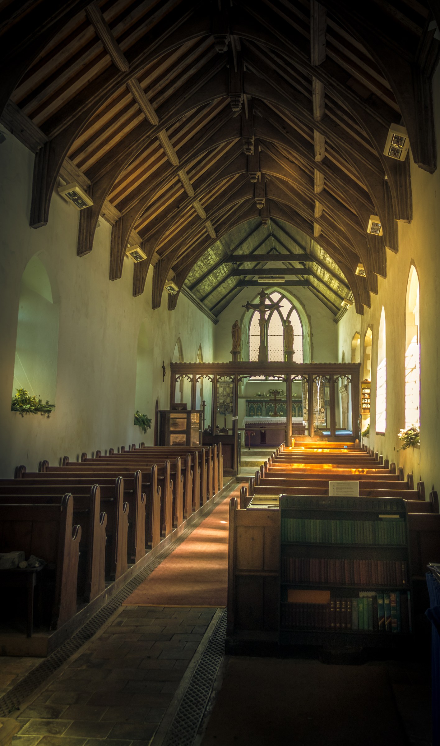 This photograph captures the serene beauty of a church interior, bathed in soft, natural light that filters through the stained glass windows, casting a space in a warm, inviting glow. The high, arched ceiling and wooden beams add a charm of traditional craftsmanship, while the rows of wooden pews lead the eye towards the altar, creating a perfect focal point. The composition, with its balanced symmetry and rich textures, evokes a sense of peace and reverence. This image is ideal for use as a living room focal piece, adding a touch of timeless elegance and spiritual ambiance to any home or office space.