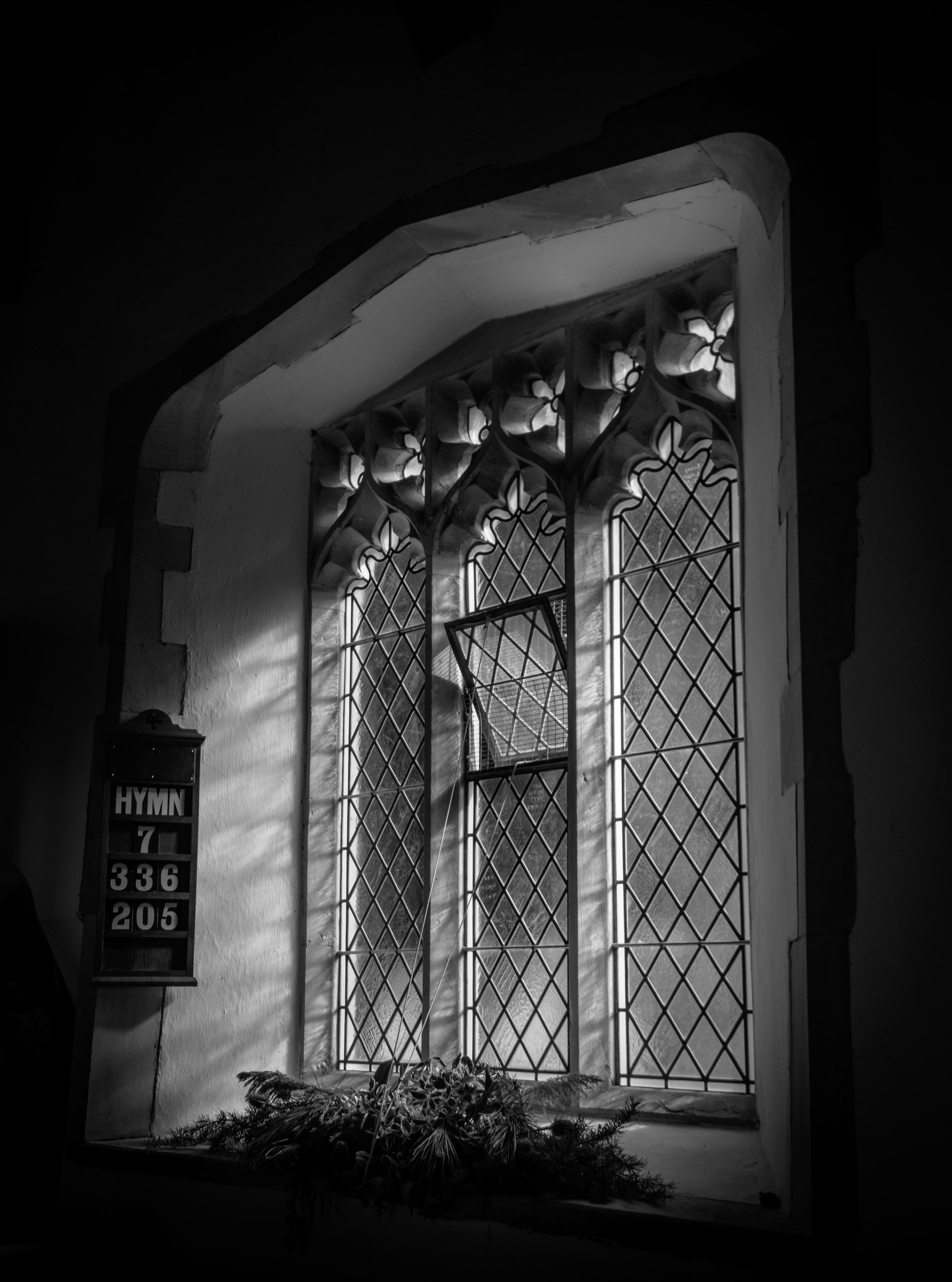 This evocative black and white photograph captures the serene beauty of a Gothic church window, bathed in soft, diffused light that filters through its intricate tracery. The interplay of light and shadow creates a dramatic and contemplative mood, highlighting the window's architectural details and the delicate patterns of the stained glass. The composition, with the hymn board in only other element in the frame, adds a perfect touch of human presence, suggesting a quiet reverence of a space. This image is ideal for wall decor in spaces that seek a ambiance of tranquility and historical elegance, such as a living room, executive office, or a modern gallery wall where it can serve as a focal piece, inspiring reflection and admiration.