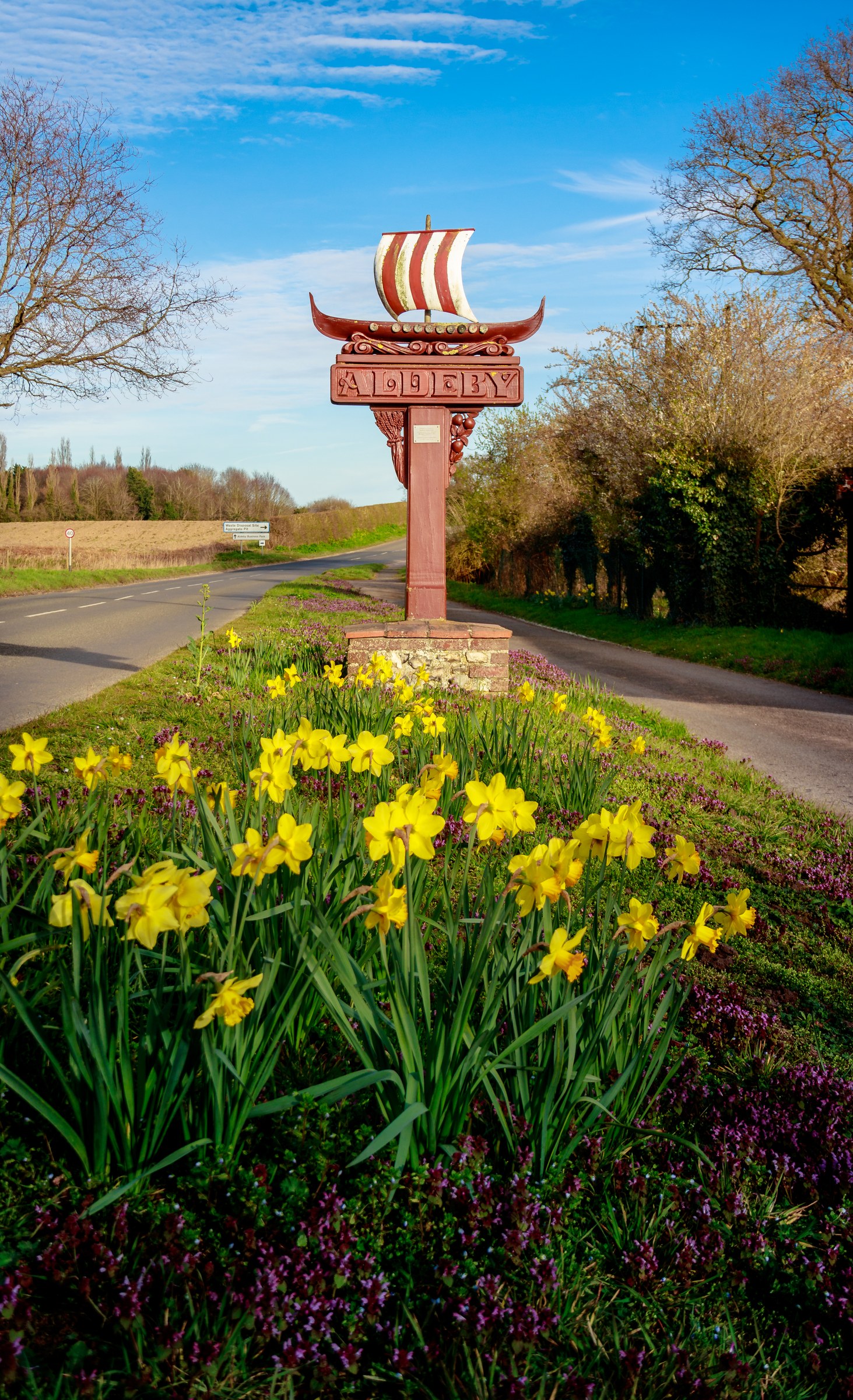 This photograph captures a serene scene featuring a Aldby Viking signpost surrounded by vibrant yellow daffodils, creating a harmonious blend of history and nature. The bright sunlight enhances the vivid colors, casting a perfect balance of light and shadow that accentuates the sign's details and the flowers' textures. The composition is balanced, with the signpost centrally positioned, drawing the viewer's eye to the rustic charm of the sign and the natural beauty of the daffodils. This image evokes a feeling of tranquility and invites viewers to imagine themselves in Viking explorers of old. This aesthetic appeal lies in its simplicity and the timeless beauty it conveys, making it an ideal focal piece for a living room or executive office, or a perfect addition to a modern gallery wall.