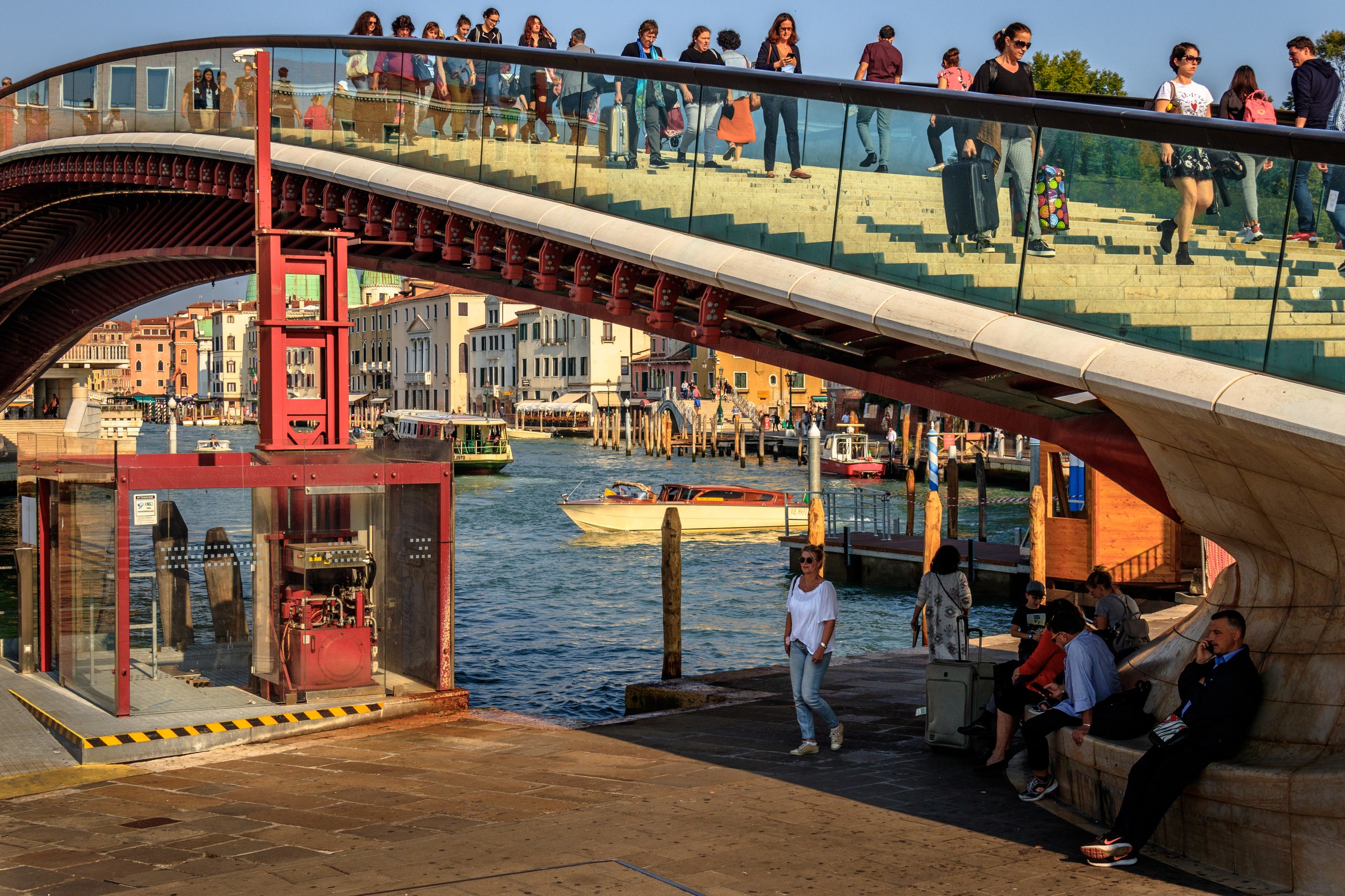 This photograph captures the enchanting essence of Venice, Italy, with its iconic red bridge and bustling waterway. The warm golden hour light bathes the scene in, highlighting the vibrant colors and textures of the bridge and the surrounding architecture. The composition beautifully balances the dynamic movement of people and boats, creating a perfect harmony of urban life and natural beauty. This image evokes a feeling of tranquility and the timeless allure of Venice, making it a ideal focal point for any living room or executive office, where it can inspire a sense of calm and elegance.