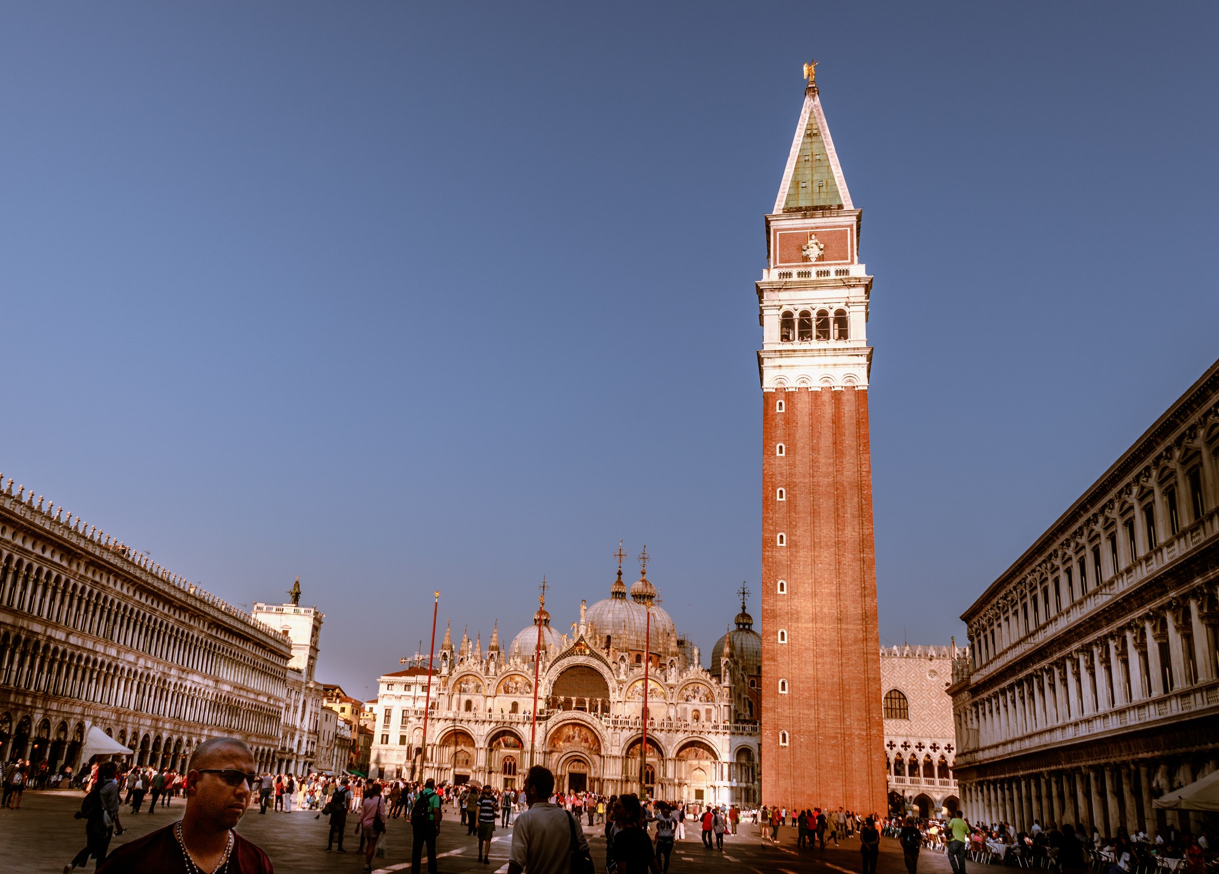 Capture the timeless beauty of St. Mark's Square in Venice with this serene and majestic photograph. The warm golden hour light bathes the iconic bell tower and the ornate basilica, creating a perfect balance of light and shadow. The composition, with the bell tower dominating the right side and the bustling square in foreground, offers a perfect blend of grandeur and everyday life. This image's aesthetic appeal lies in its rich colors and the sense of history it evokes, making it a perfect focal piece for a living room or an executive office. Hang it in a corner of your modern gallery wall to inspire and remind you of the beauty of the world.
