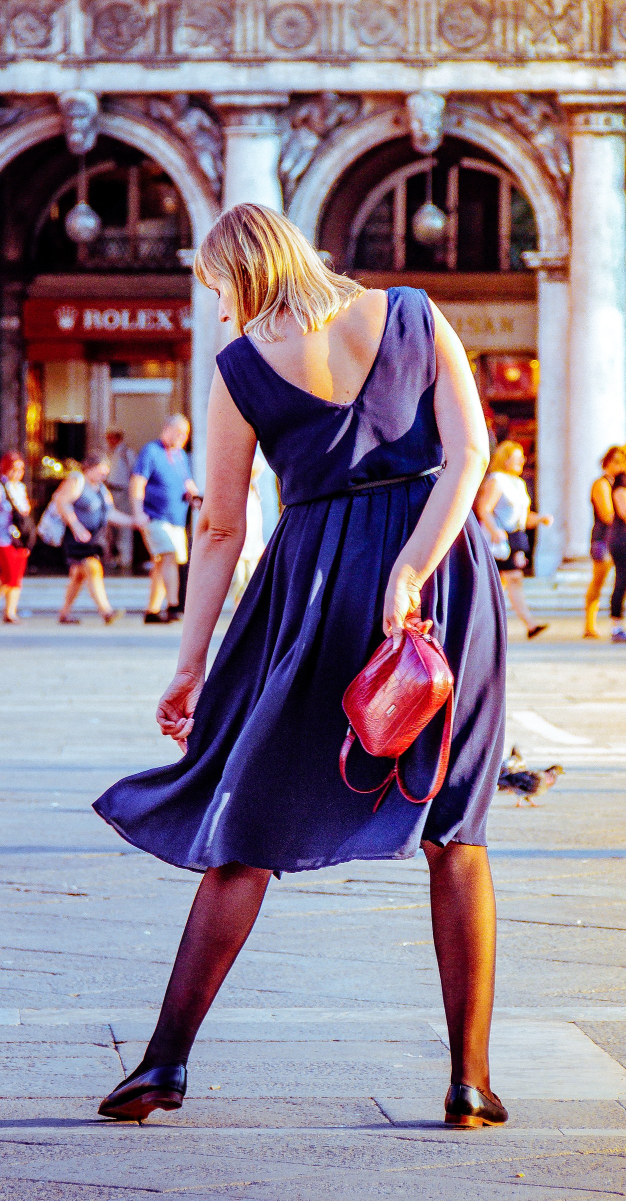 This photograph captures a essence of timeless elegance, set against the vibrant backdrop of Venice's iconic Piazza San Marco. The soft, golden hour lighting bathes the scene in in a warm glow of sunset, highlighting the woman's flowing navy blue dress and the rich texture of her red leather purse. The composition, with the subject walking away, invites the viewer to imagine the story behind her journey. This image's aesthetic appeal lies in its balance of color, movement, and cultural context, making it a perfect focal piece for a living room or a statement wall in an executive office. The image evokes a feeling of adventure and sophistication, perfect for those who appreciate the beauty of travel and fine art.