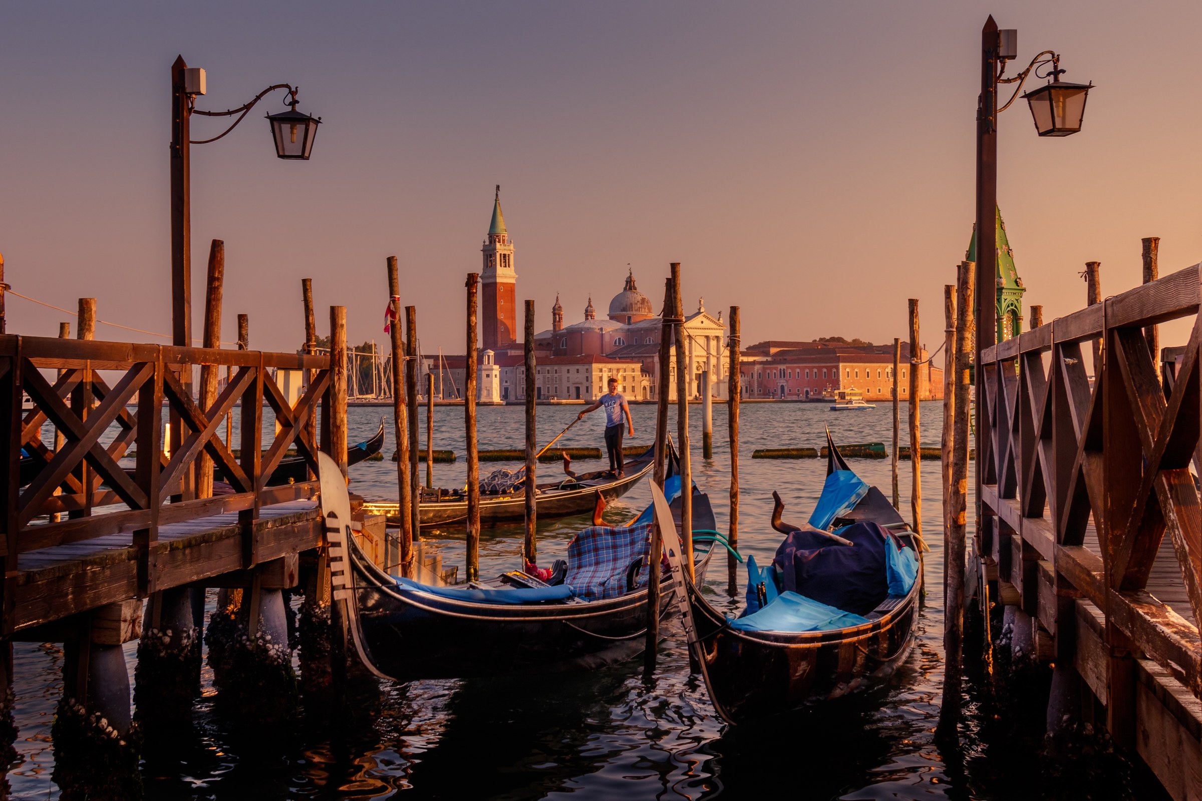 This photograph captures the enchanting essence of Venice at sunset, with gondolas gently bobbing on the calm waters. The warm hues of the setting sun cast a scene in a soft glow of golden light, creating a tranquil and romantic atmosphere. The composition beautifully frames the iconic gondolas against the backdrop of the historic San Giorgio Maggiore church, adding a perfect balance of natural and architectural beauty. This rich, warm tones and the serene setting make this image a perfect piece for a living room, where it can serve as a focal point, evoking the feeling of a peaceful evening in Venice.
