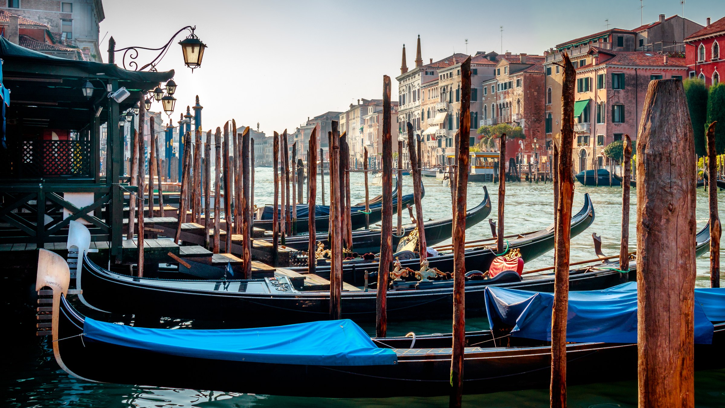 Capture the essence of Venice with this stunning photograph of gondolas moored along the Grand Canal. The warm, golden hour light bathes scene in in a soft glow of sunset, creating a perfect balance of tranquility and romance. The composition, with its leading lines and symmetrical arrangement of gondolas, draws the viewer's eye across the tranquil water, evoking a calm and timeless beauty of this iconic city. This rich, deep blues of the gondolas' tarps contrast beautifully with the warm tones of the buildings and water, making this image a perfect focal point for any living room or executive office, where it can inspire a soul and add a charm of Venice to your space.