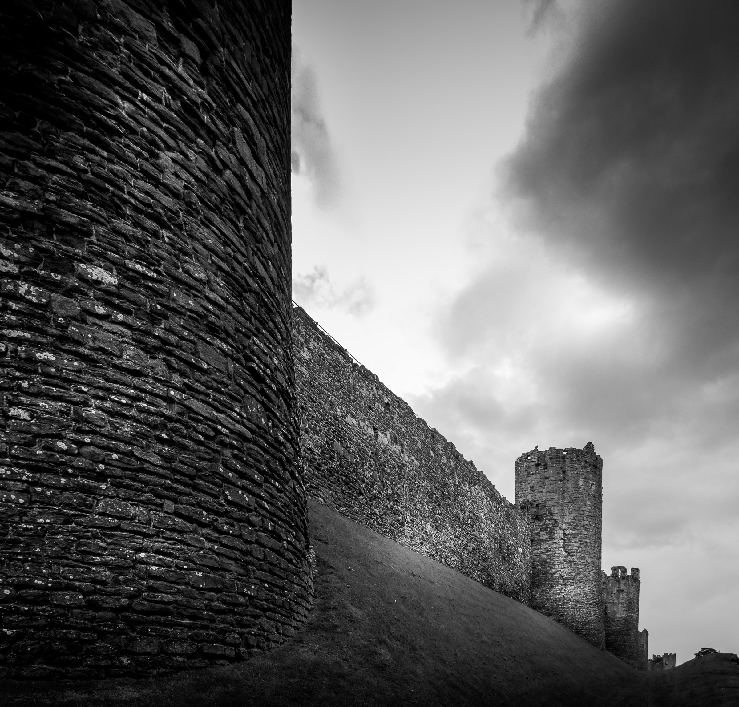 This striking black and white photograph captures the timeless beauty of ancient castle walls, evoking a grandeur and mystery of medieval times. The dramatic interplay of light and shadow, combined with the rugged texture of the stone, creates a perfect balance of ruggedness and elegance. The composition, with its strong vertical lines and the imposing tower in the distance, draws the viewer's eye deep into the scene, making it a perfect focal point for any modern or traditional interior. Whether displayed in living room or an executive office, this piece adds a perfect touch of historical sophistication and artistic flair.