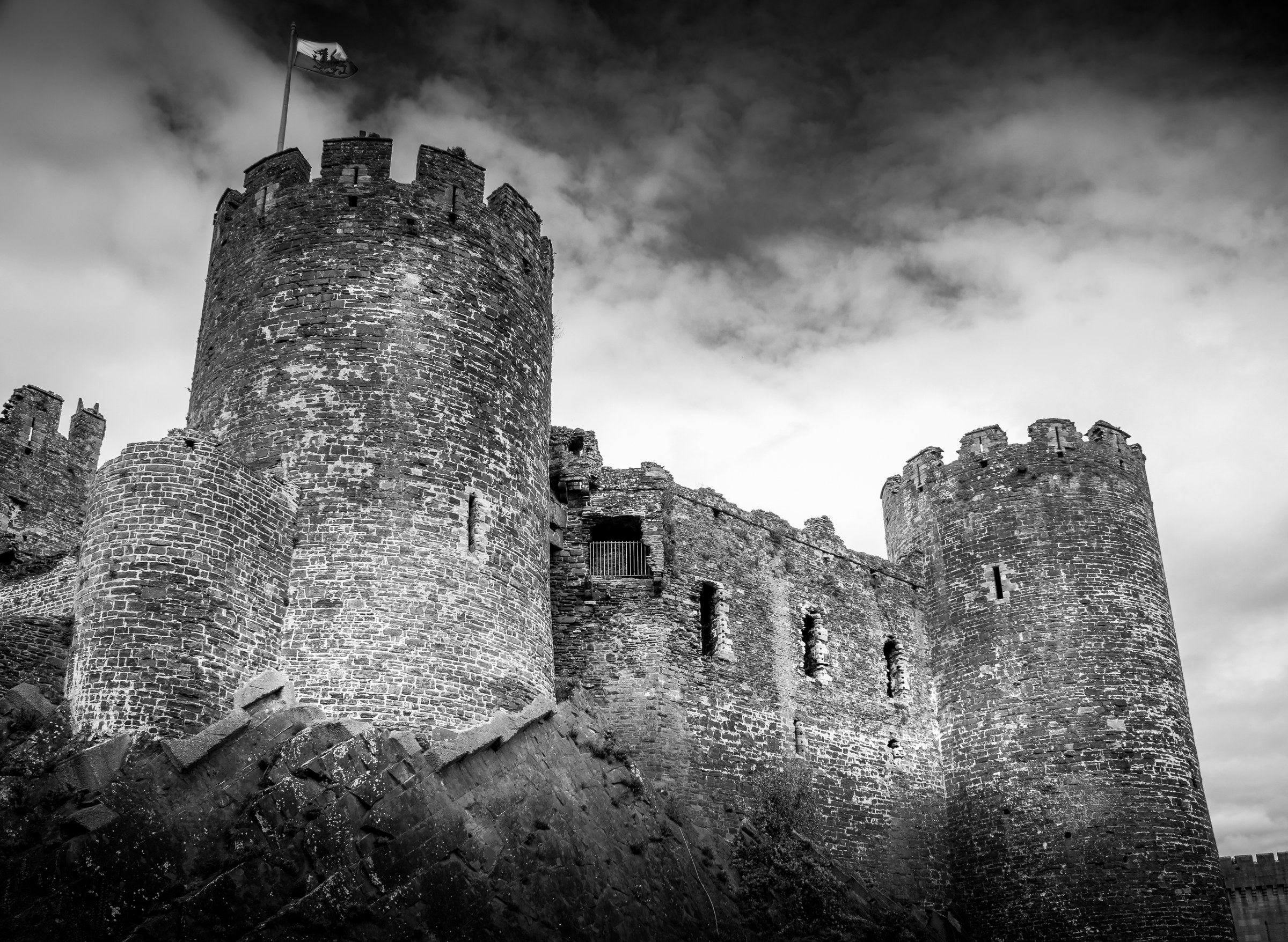 This striking black and white photograph captures the timeless grandeur of a historic castle, its rugged stone walls and crenellated towers standing as a testament to medieval architecture. The dramatic interplay of light and shadow, accentuated by the overcast sky, evokes a mood of mystery and historical depth. The composition, with its strong vertical lines and symmetrical balance, creates a perfect focal point for any modern or traditional interior. Ideal for a living room as a dramatic statement piece or an executive office to inspire a spirit of leadership and resilience.