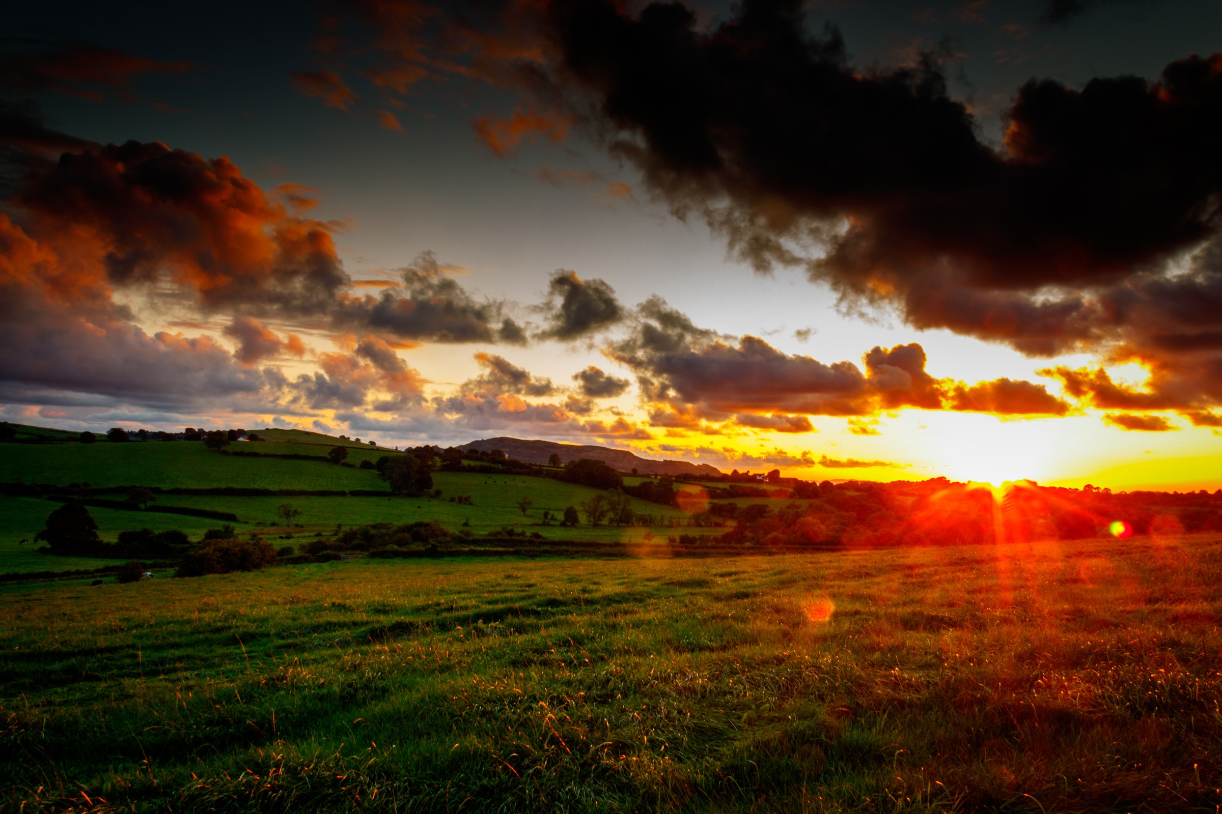 This breathtaking photograph captures the serene beauty of a sunset over lush, rolling hills, bathed in the warm, golden hues of the setting sun. The dramatic interplay of light and shadow, combined with the rich, vibrant colors, evokes a feeling of tranquility and awe. The composition, with the sun positioned low on the horizon, creates a perfect focal point, drawing the viewer's eye across the expansive landscape. This image's aesthetic appeal lies in its ability to transport viewers to a peaceful solitude of nature, making it an ideal choice for wall decor in living room, executive office, or modern gallery wall. Hang it as a centerpiece of a room to inspire calm and reflect the beauty of the natural world.