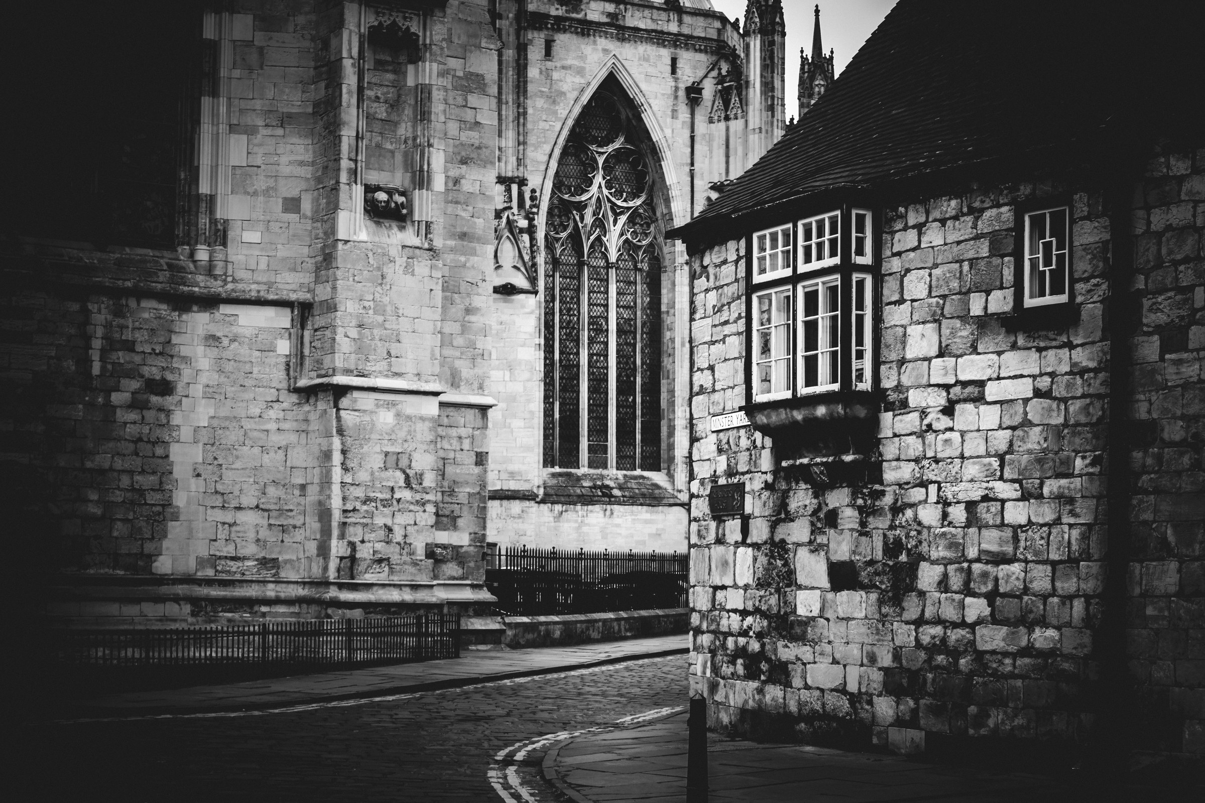 This evocative black and white photograph captures the timeless beauty of an urban Gothic setting, where the interplay of light and shadow creates a illusion of a sun setting behind the ancient stone walls. The composition, with its curved street leading the eye towards the cathedral's intricate window, evokes a feeling of a past merging with the present. The aesthetic appeal lies in its stark contrast and the dramatic interplay of light and shadow, making it a perfect focal piece for a living room or a executive office, where it can adds a perfect touch of sophistication and historical charm.